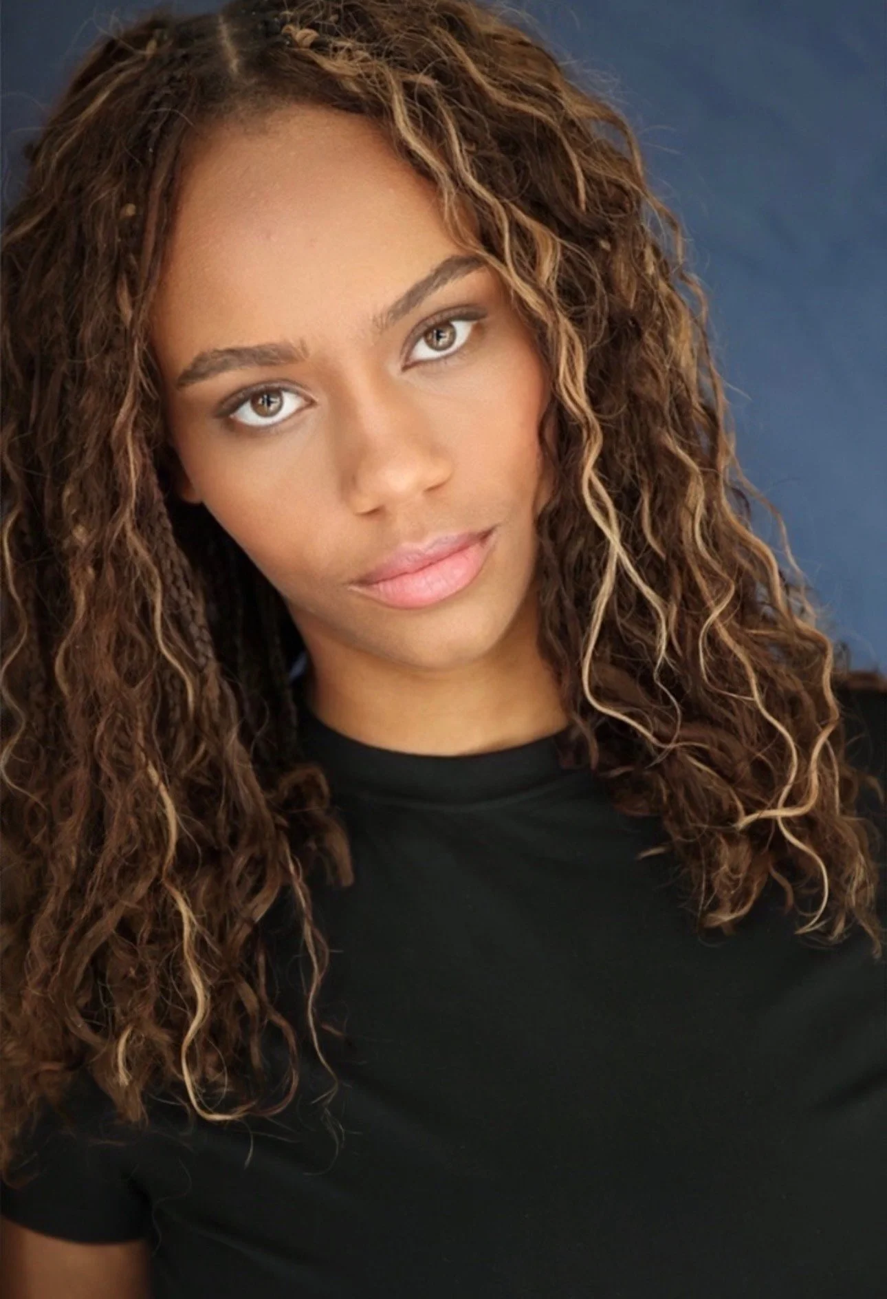 Close-up portrait of a young woman with long, curly, light brown hair and brown eyes, wearing a black top, against a dark background.