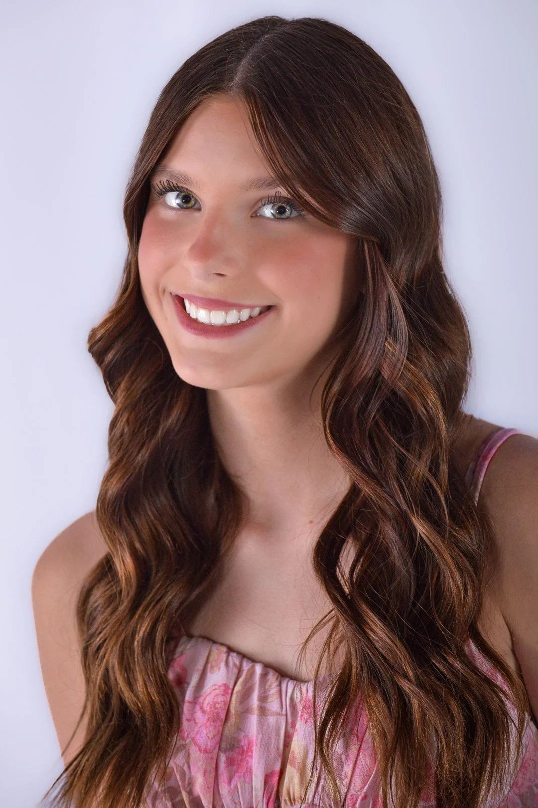 Close-up of a young woman with long wavy brown hair wearing a pink floral dress and smiling at the camera.