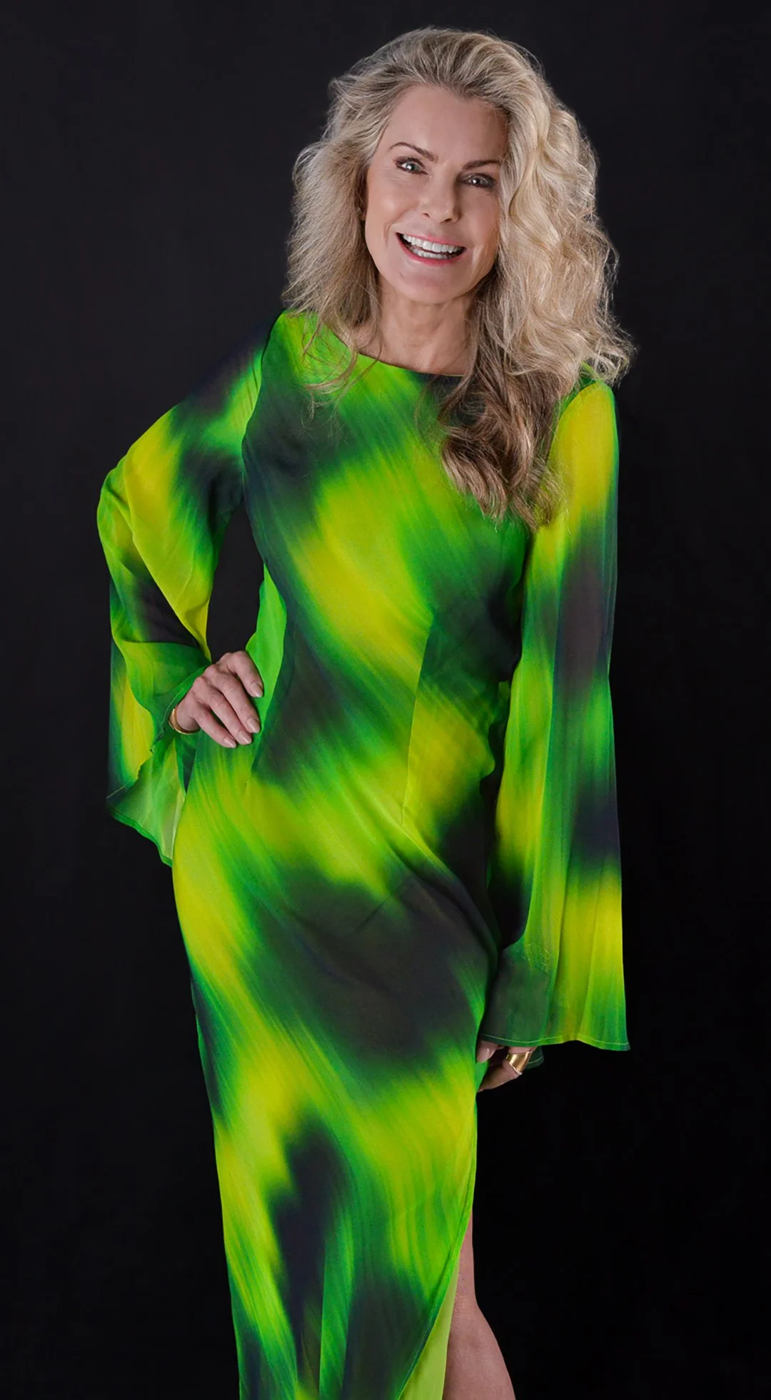 A woman with blonde, curly hair wearing a vibrant green and black flowing dress, smiling and standing against a black background.