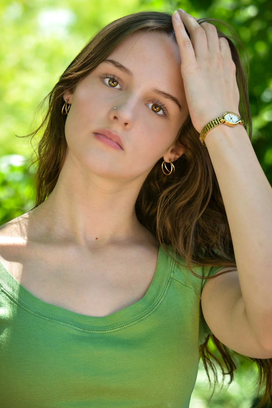 A young woman with long brown hair, light skin, and hazel eyes, wearing a green top, gold earrings, and a gold watch, standing outdoors with a blurred green leafy background, touching her hair on her forehead with her right hand.