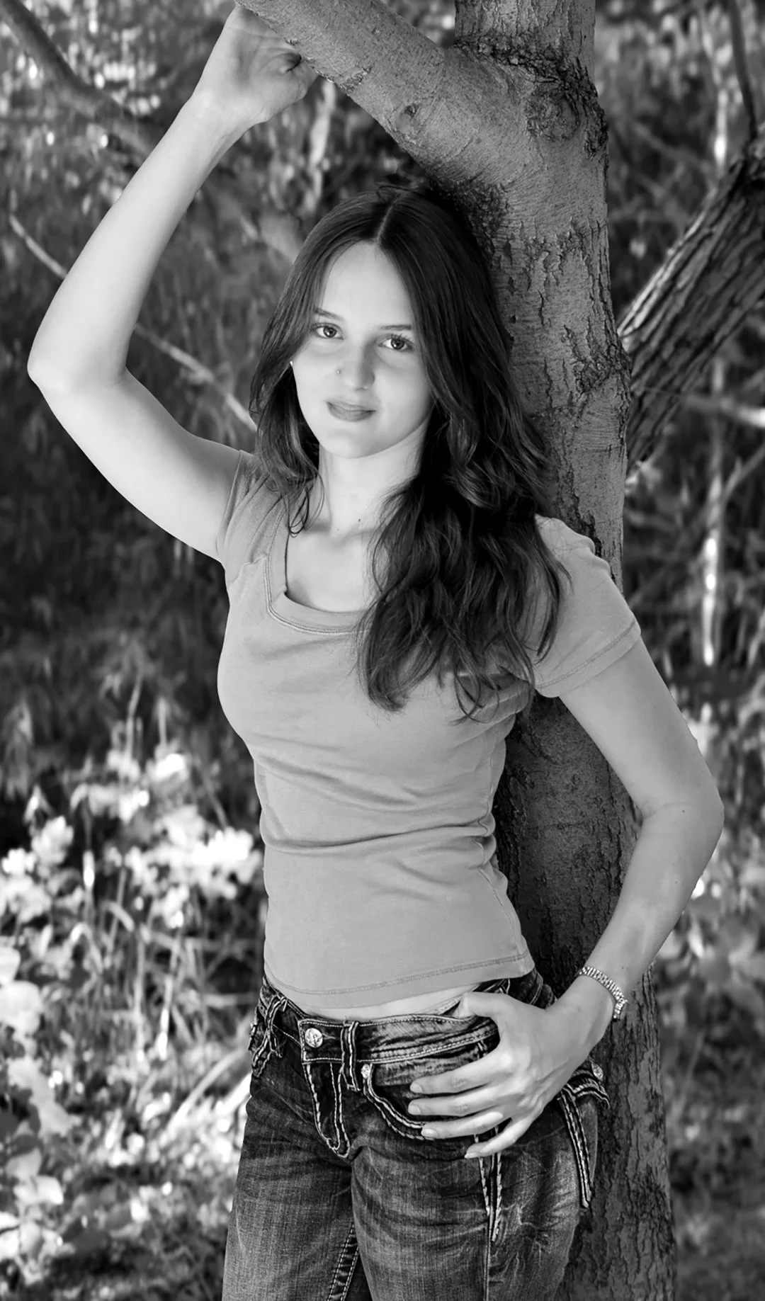 Young woman with long wavy hair leaning against a tree in an outdoor setting, wearing a fitted t-shirt and jeans.