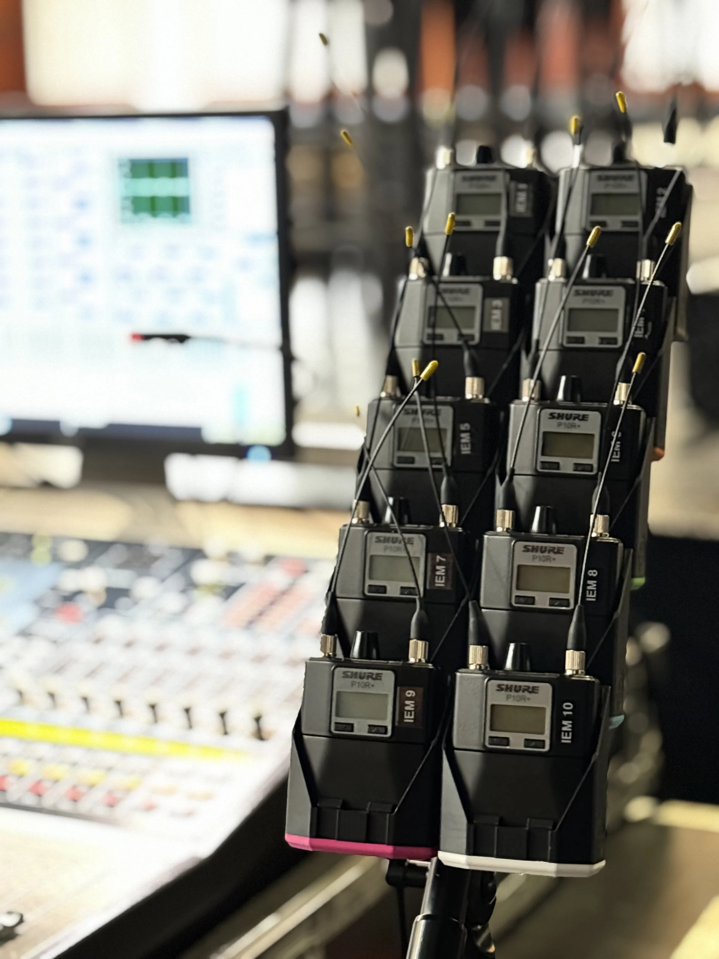 A rack of multiple wireless microphone receivers with small antennas, set up near a mixing console and a computer monitor.