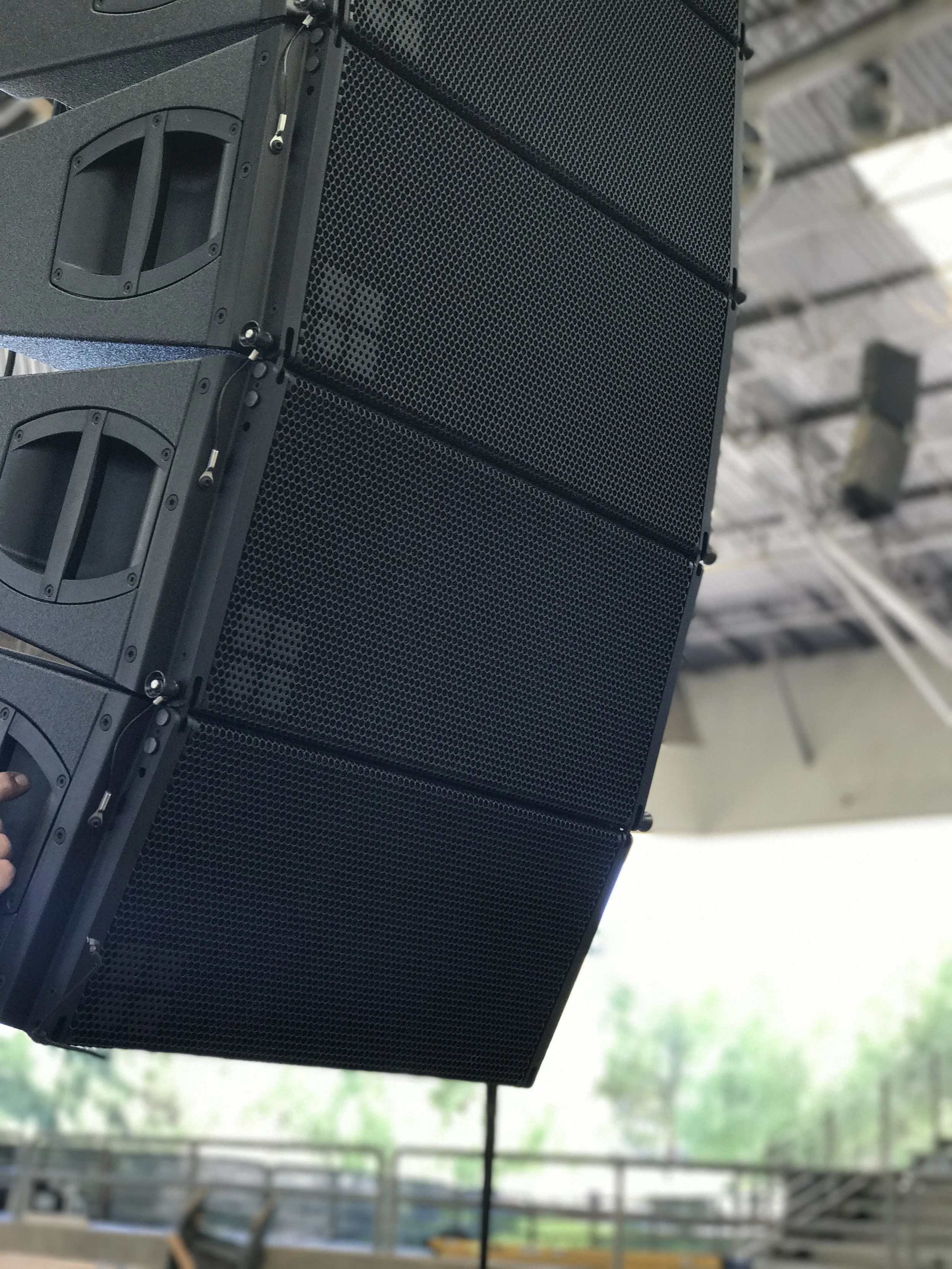 Large black speaker system with multiple speaker units mounted on a stage rigging in an indoor venue.