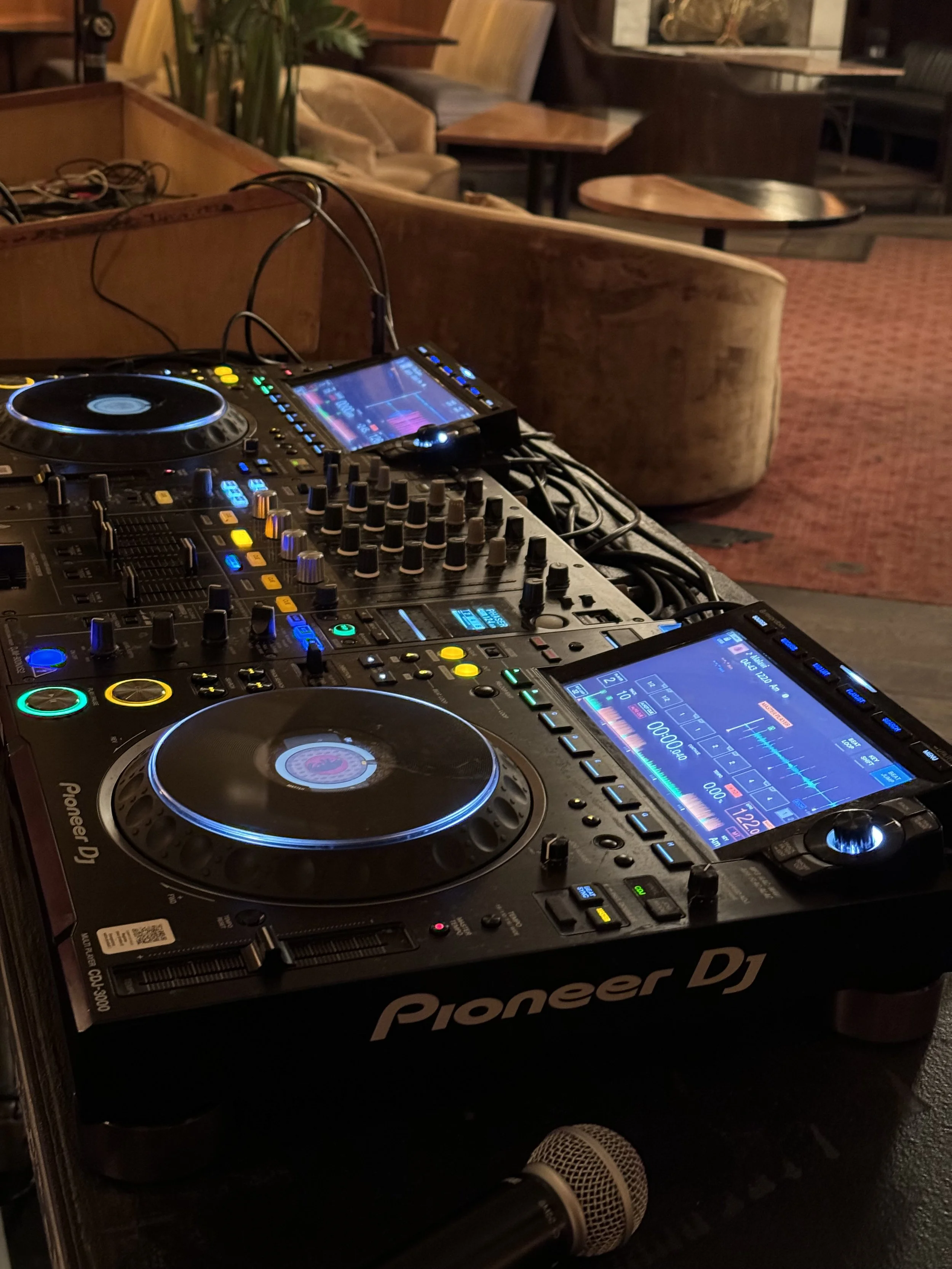 DJ equipment, including Pioneer DJ controllers with colorful lights, set up on a table in a cozy lounge with couches and tables in the background.