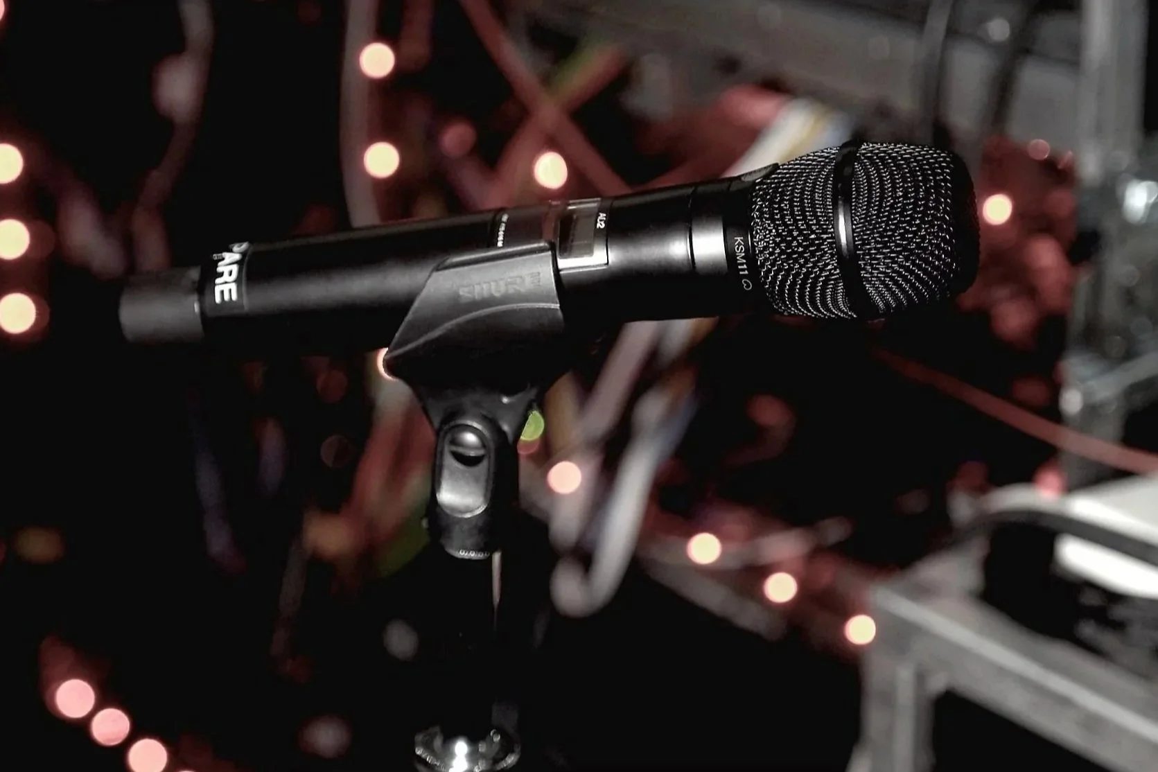 A microphone on a stand in front of blurred string lights and metallic structure in a dark setting.