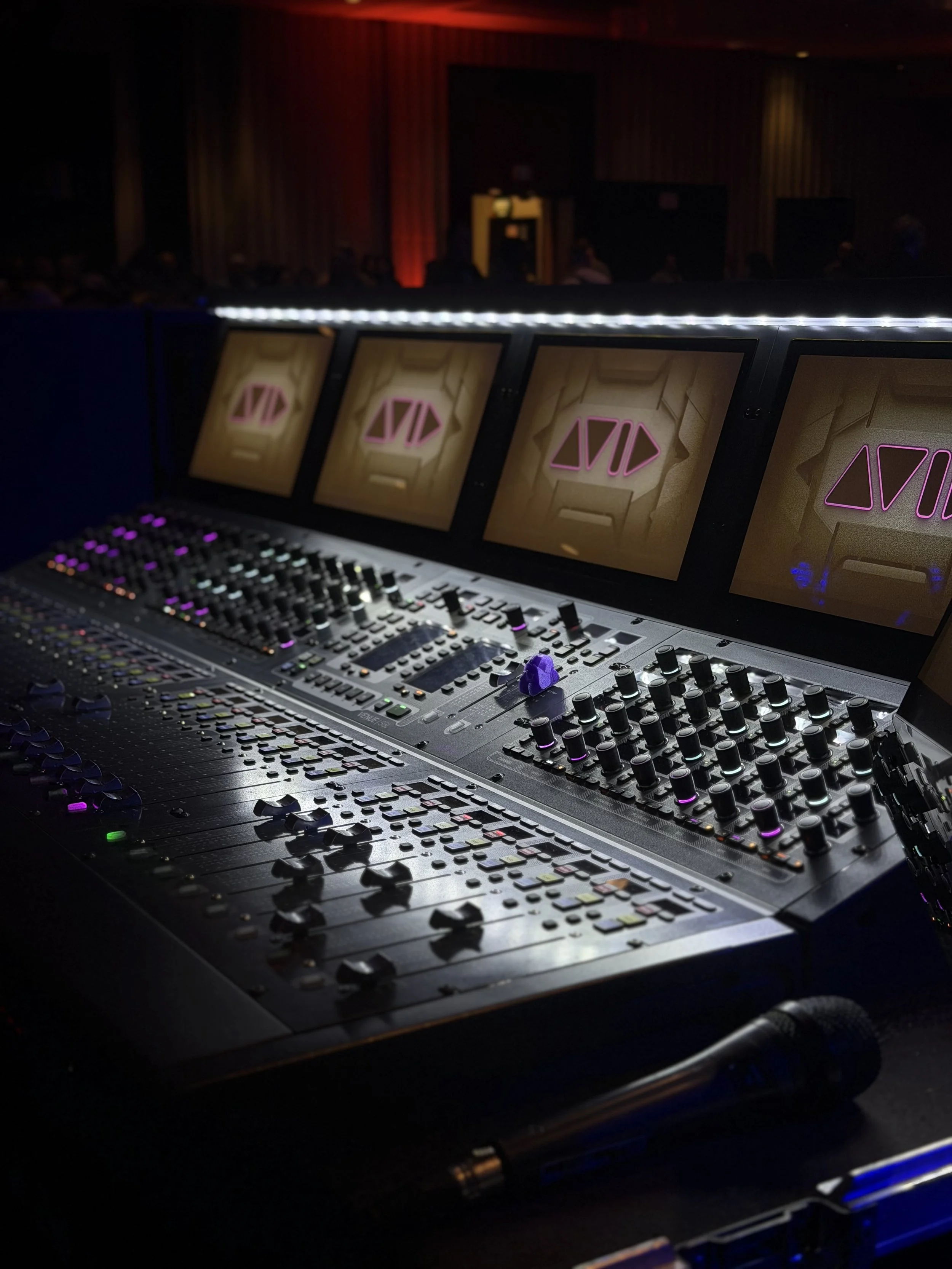 Close-up of a professional audio mixing console with multiple screens displaying a geometric logo, set in a dark, stage-like environment.
