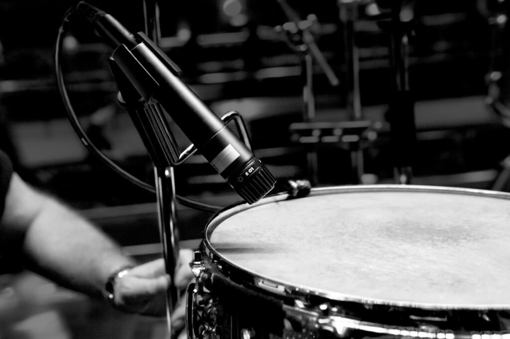 A close-up black and white photo of a microphone positioned above a snare drum, with a person's hand and arm visible on the left side of the image, in a dimly lit recording studio or stage setting.