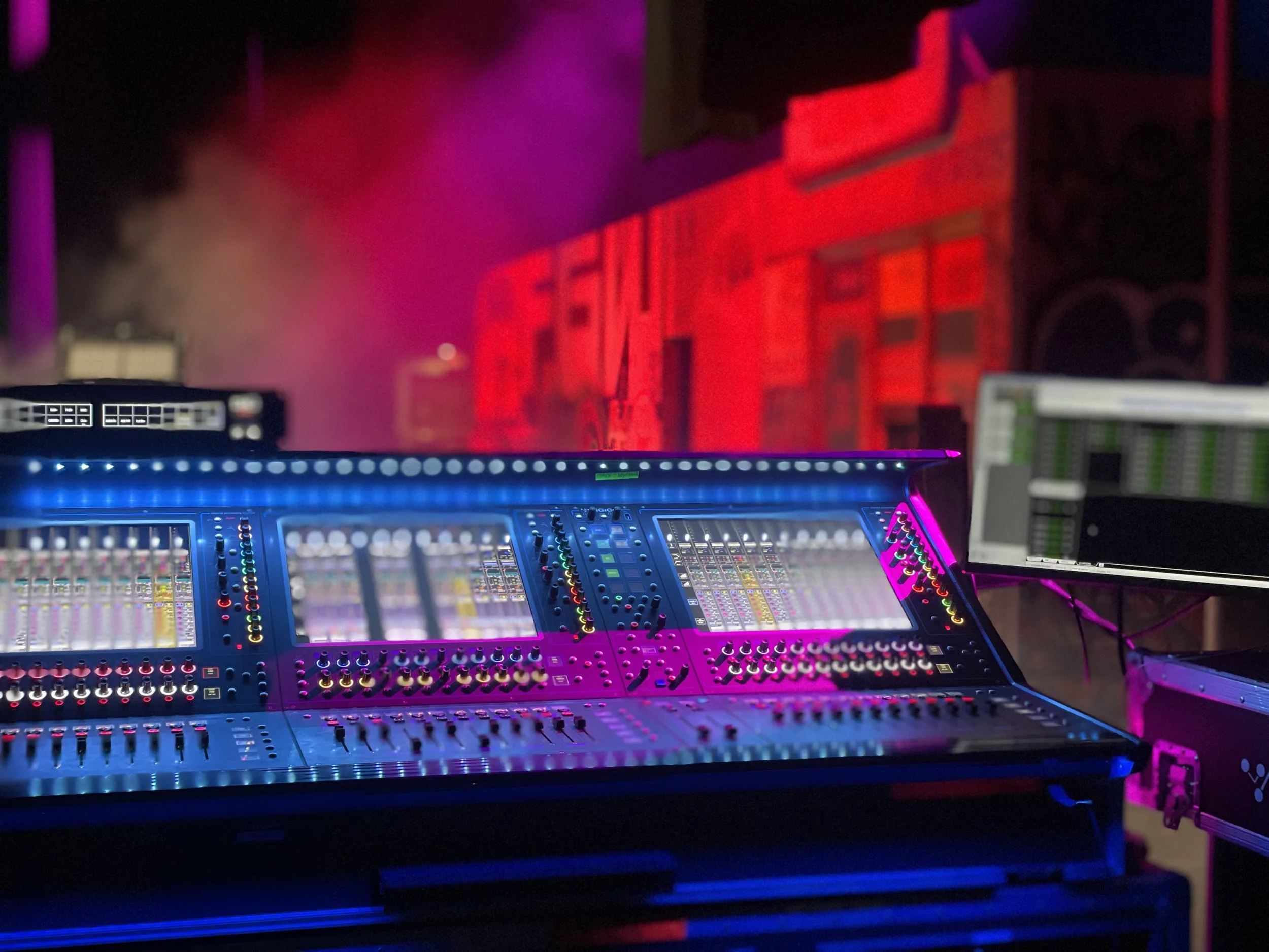 Close-up of a digital audio mixing console with colorful lights, situated in a dimly lit stage or studio with red and purple lighting.