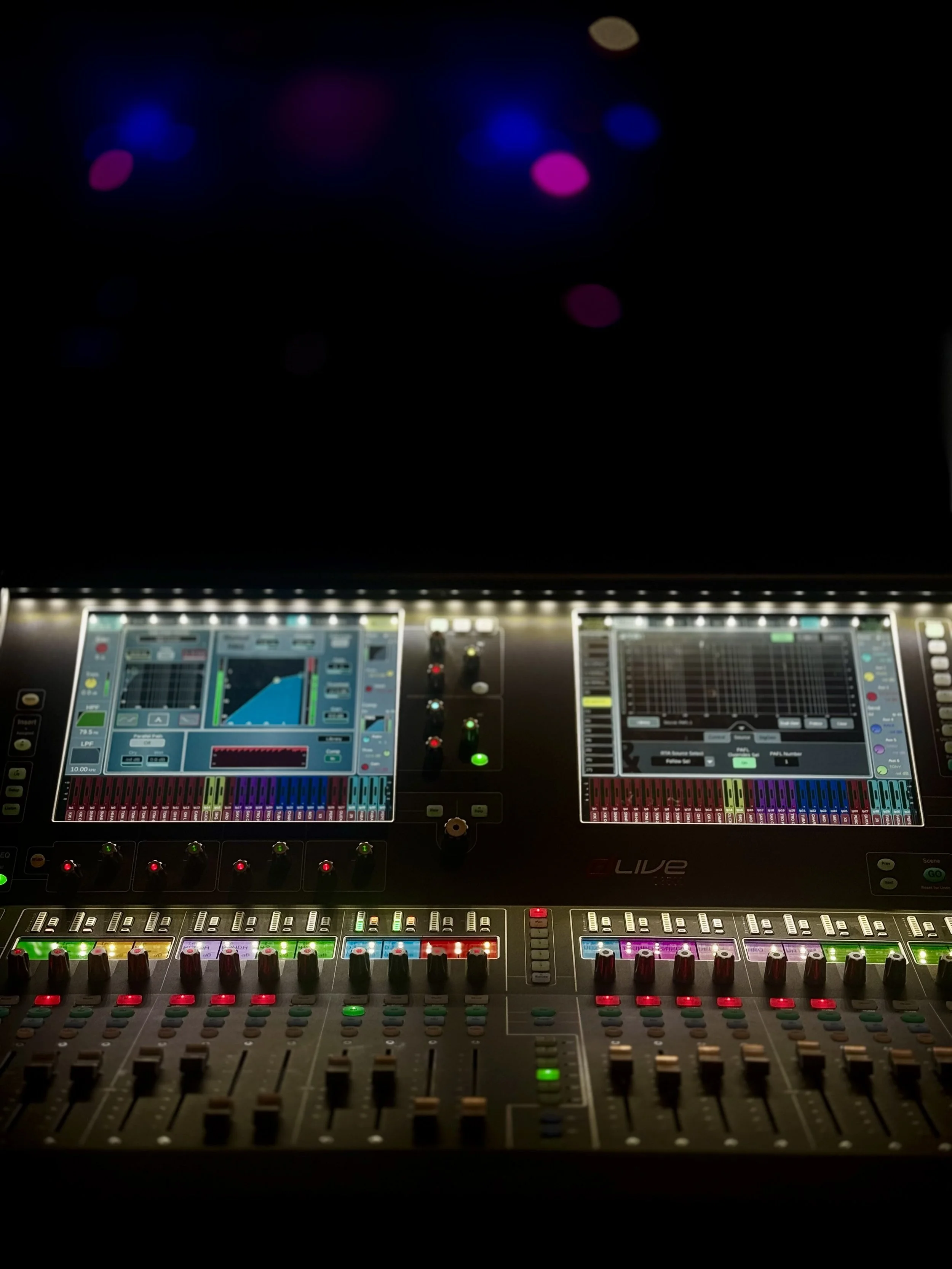 An Allen & Heath C3500 digital sound mixing console with various colorful sliders, knobs, and digital screens showing equalizer and sound level graphs, illuminated in a dimly lit environment with blurred colorful lights in the background.