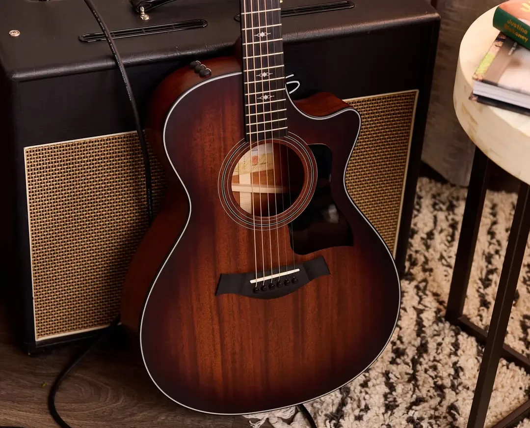 An acoustic guitar leaning against a black amplifier, with a small white table nearby holding books and other items. The guitar has a dark wood finish with white trim and is on a patterned rug.