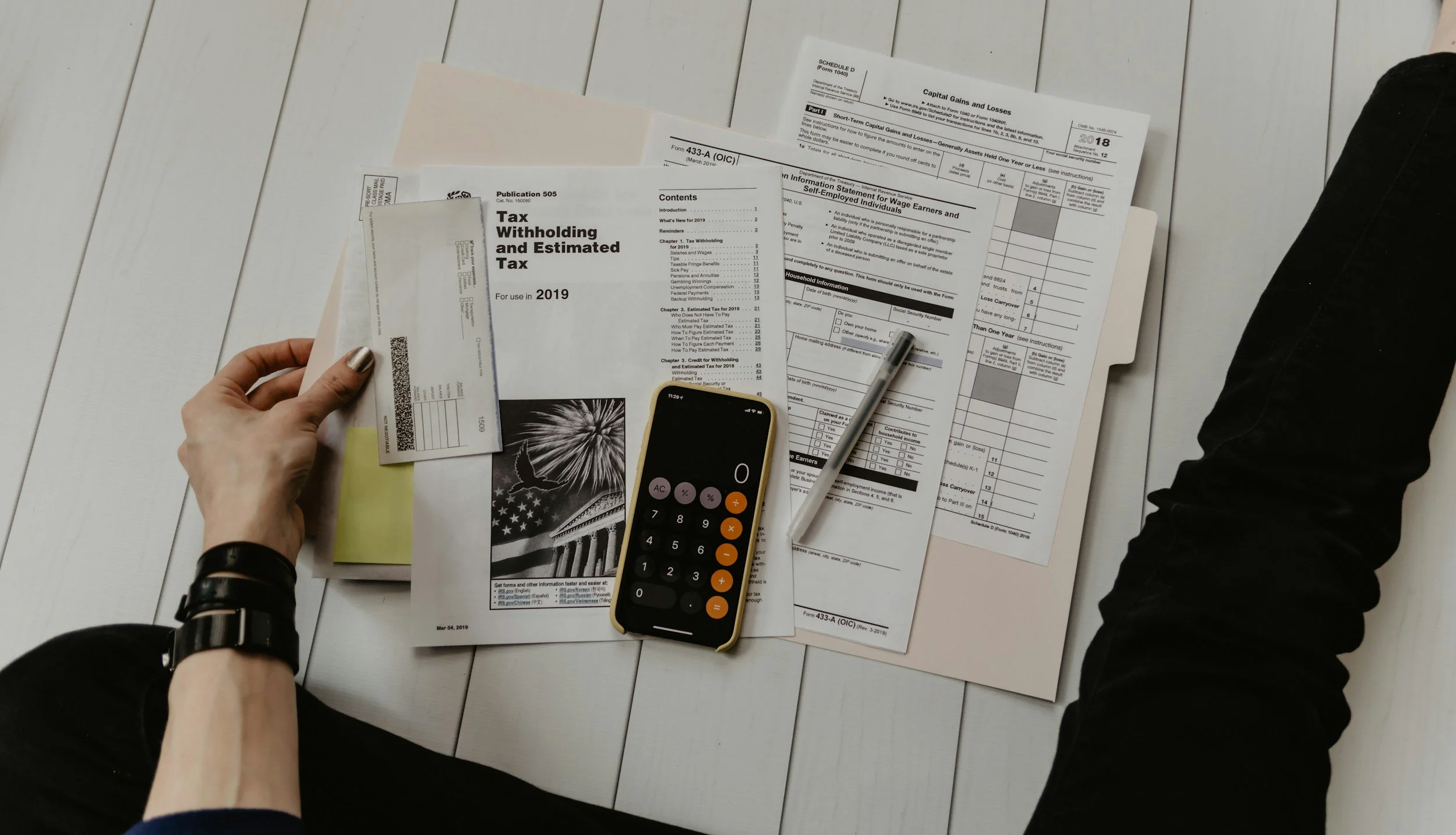 A person working on tax documents with a calculator, phone, and various forms on a white wooden table.