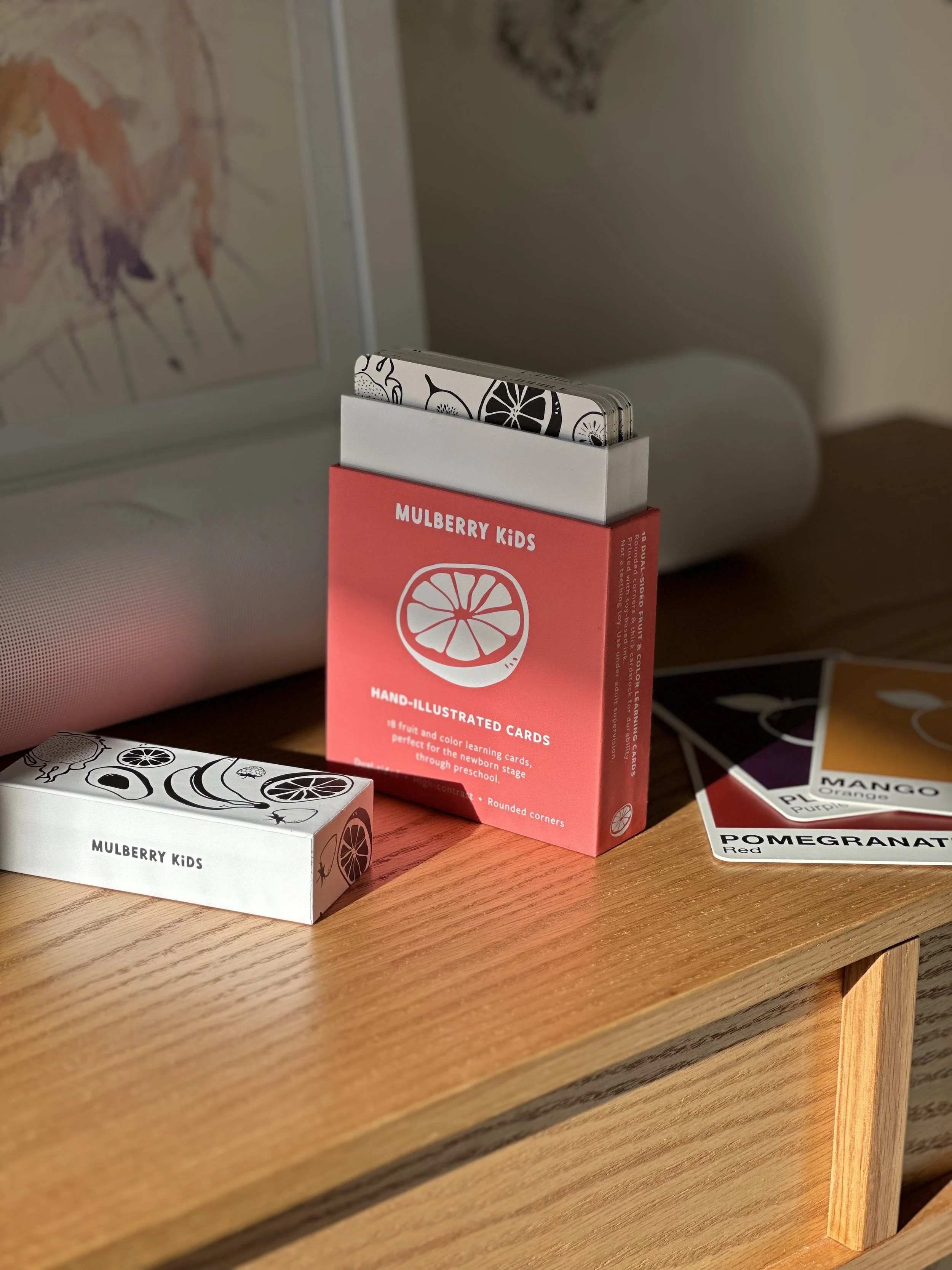A set of Mulberry Kids fruit and color learning cards, featuring illustrations of various fruits, on a wooden surface next to a box and some cards.