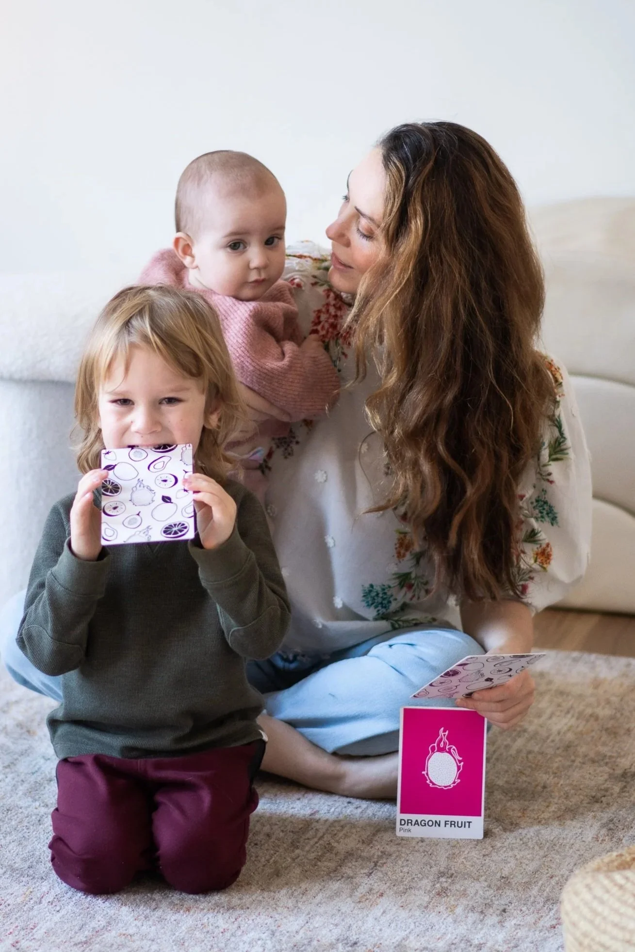 A woman with long brown hair playing with two young children in a living room. One child, a girl with blonde hair, is kneeling on a carpet holding a card, and the other child, a baby in pink, is being held by the woman.