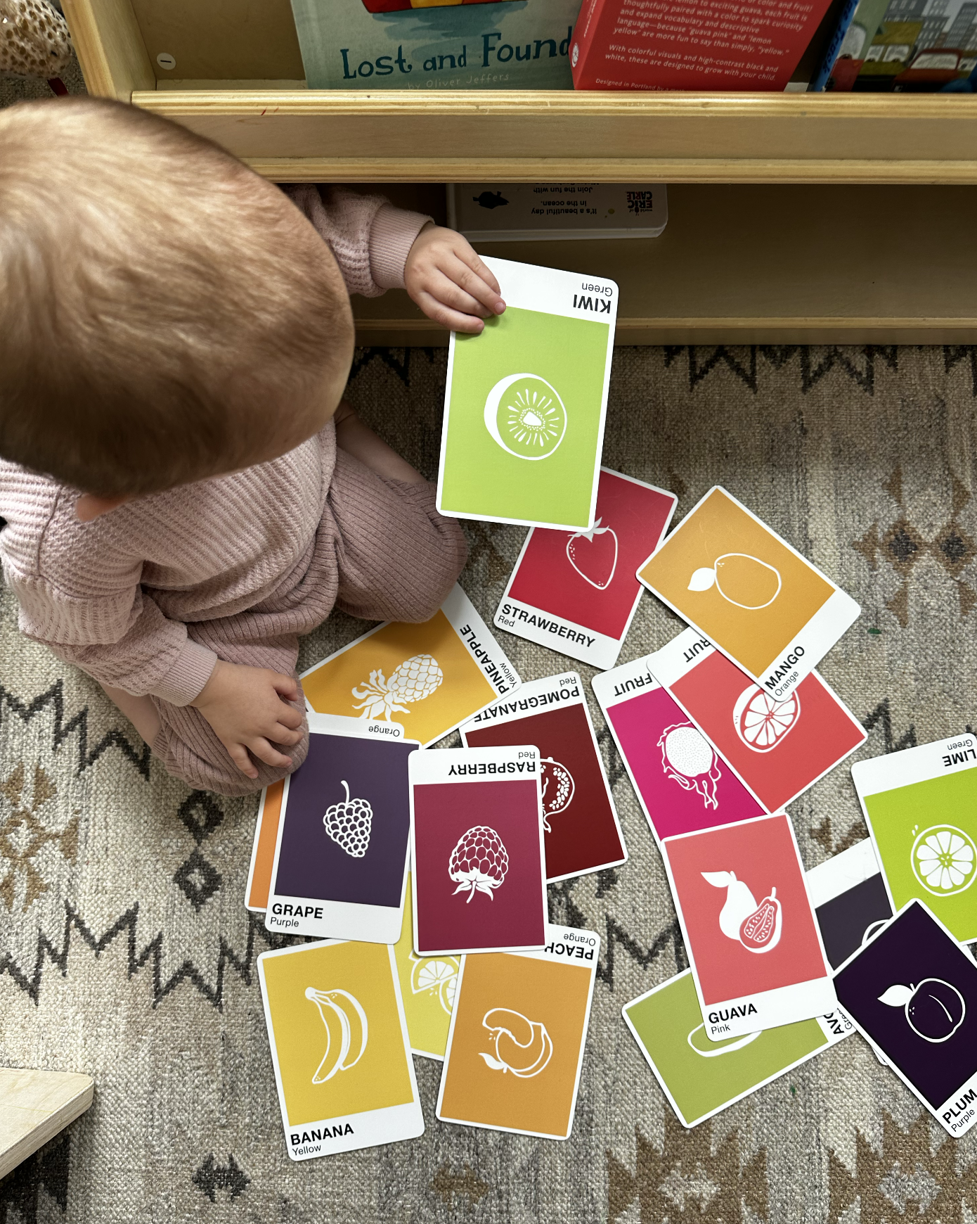 A young child sitting on a patterned rug, playing with colorful fruit and berry flashcards. The cards include images and names of various fruits such as a banana, grape, strawberry, mango, guava, and plum, with some cards face-up and others face-down.