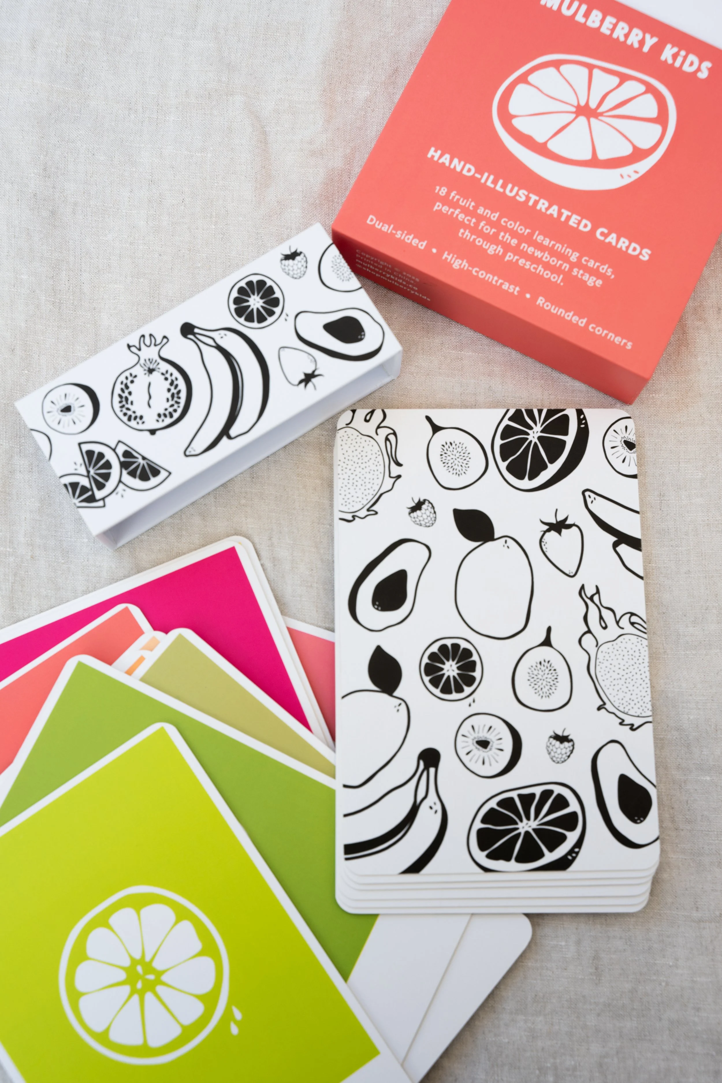 The image shows a set of fruit-themed learning cards, including a black-and-white card with various fruit illustrations and a colorful card deck with vibrant fruit images. There's also an orange-red box labeled "Mulberry Kids Hand-Illustrated Cards" containing 18 dual-sided, high-contrast, rounded-corner cards designed for preschool learning.