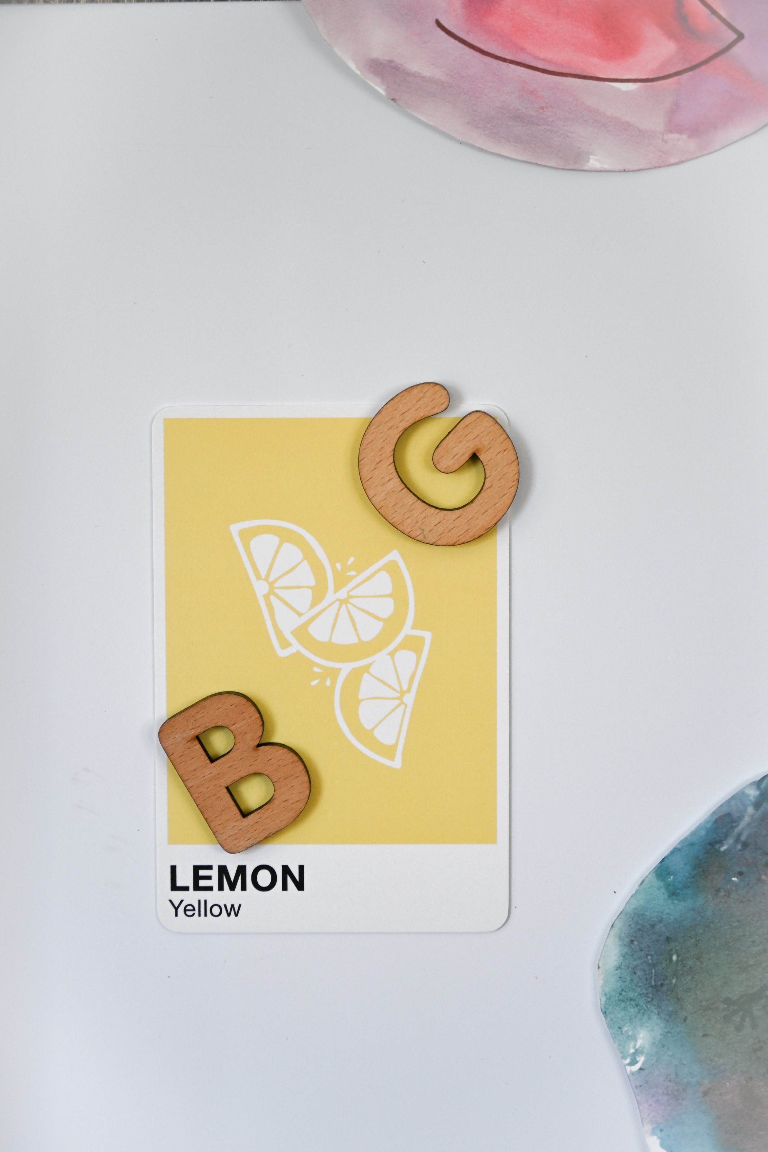 A color identification card for lemon, yellow in color, with white illustrations of lemon slices, on a white surface. There are two wooden letter and shape pieces, with a 'B' letter at the bottom left and a spiral shape at the top right.