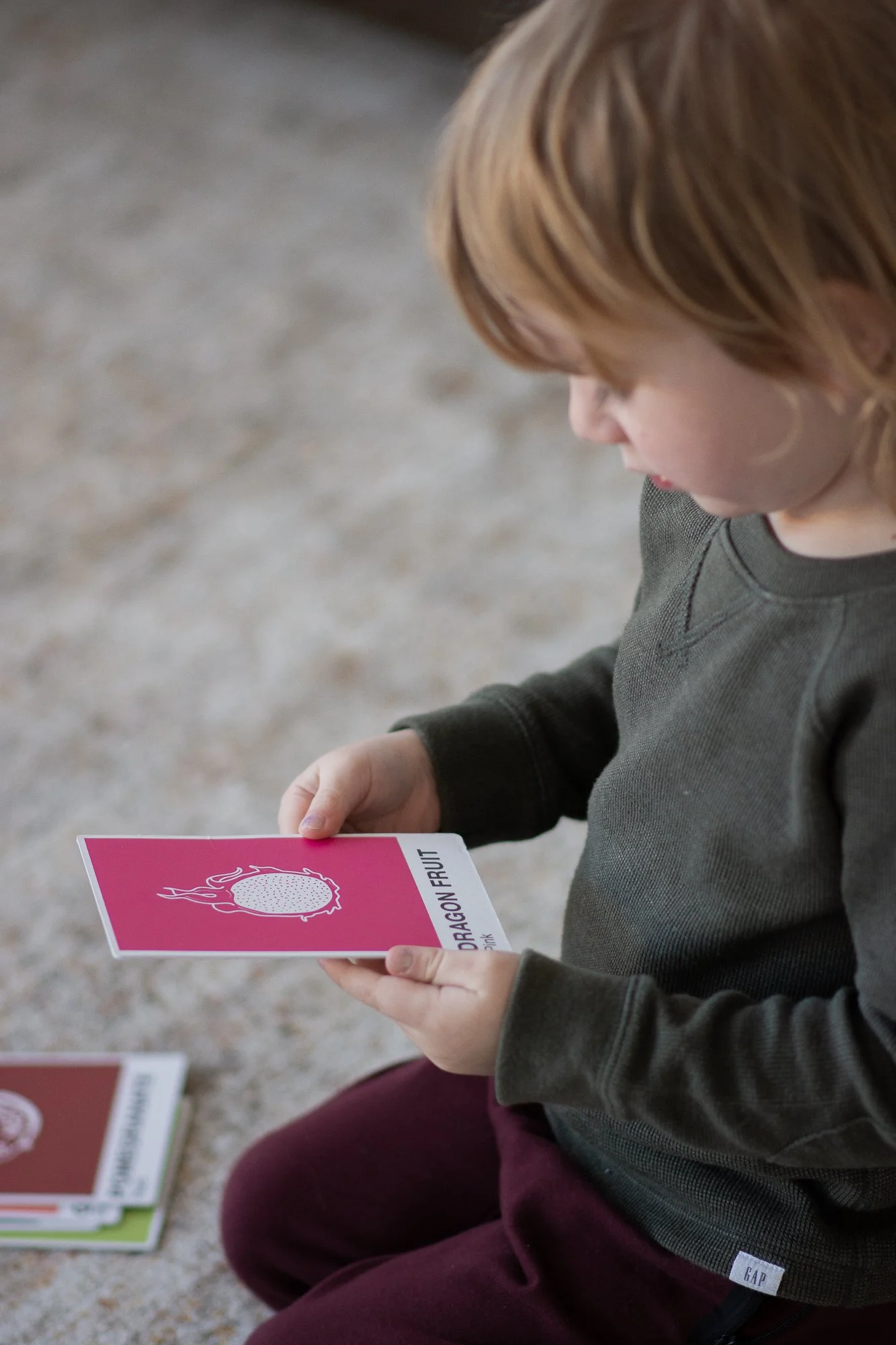 A young child with red hair wearing a dark green sweatshirt and maroon pants is sitting on a beige carpet, holding a pink flashcard with a dragon fruit illustration and the text 'DRAGON FRUIT'. Additional flashcards are on the floor next to them.