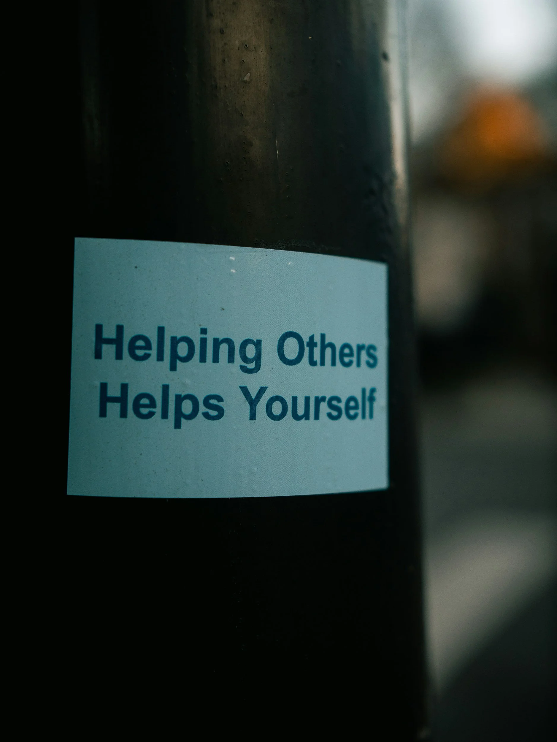 A sticker on a dark post or pole that reads 'Helping Others Helps Yourself' in blue text.