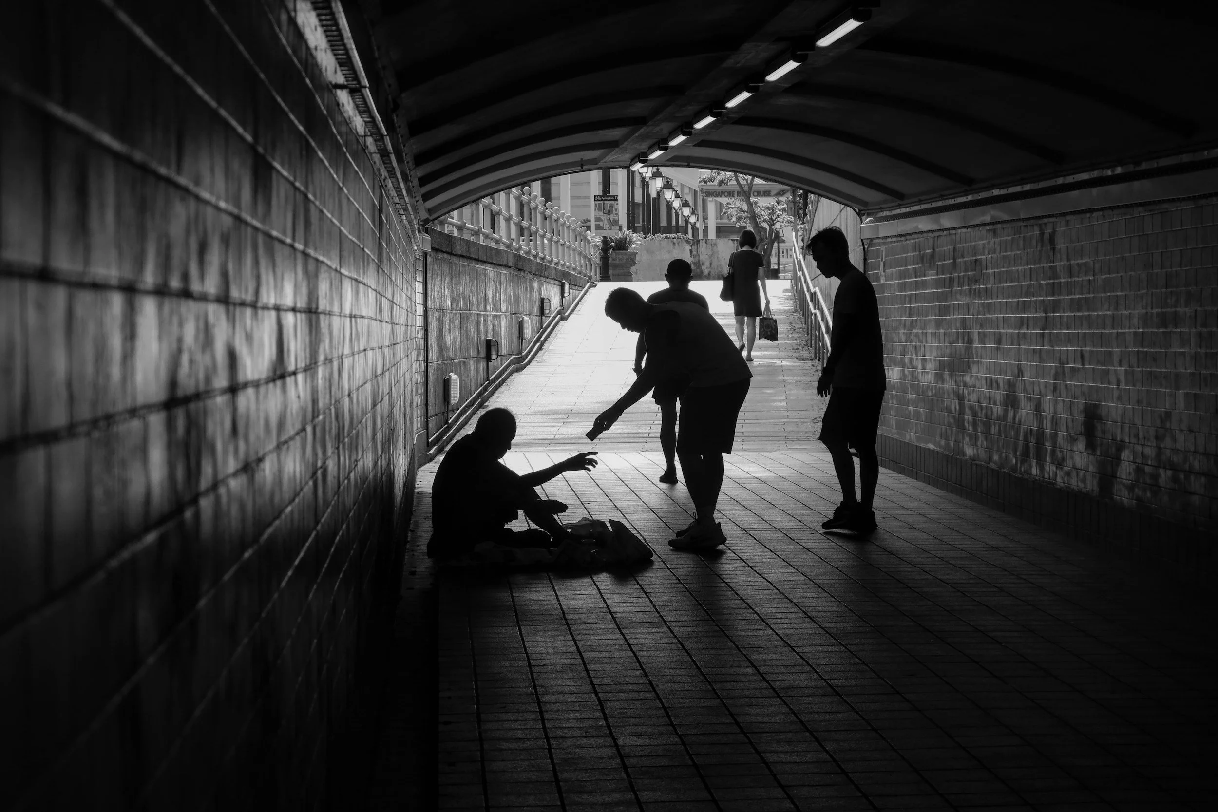 Silhouettes of people under a bridge, with one person sitting on the ground and others standing around, in an urban setting.