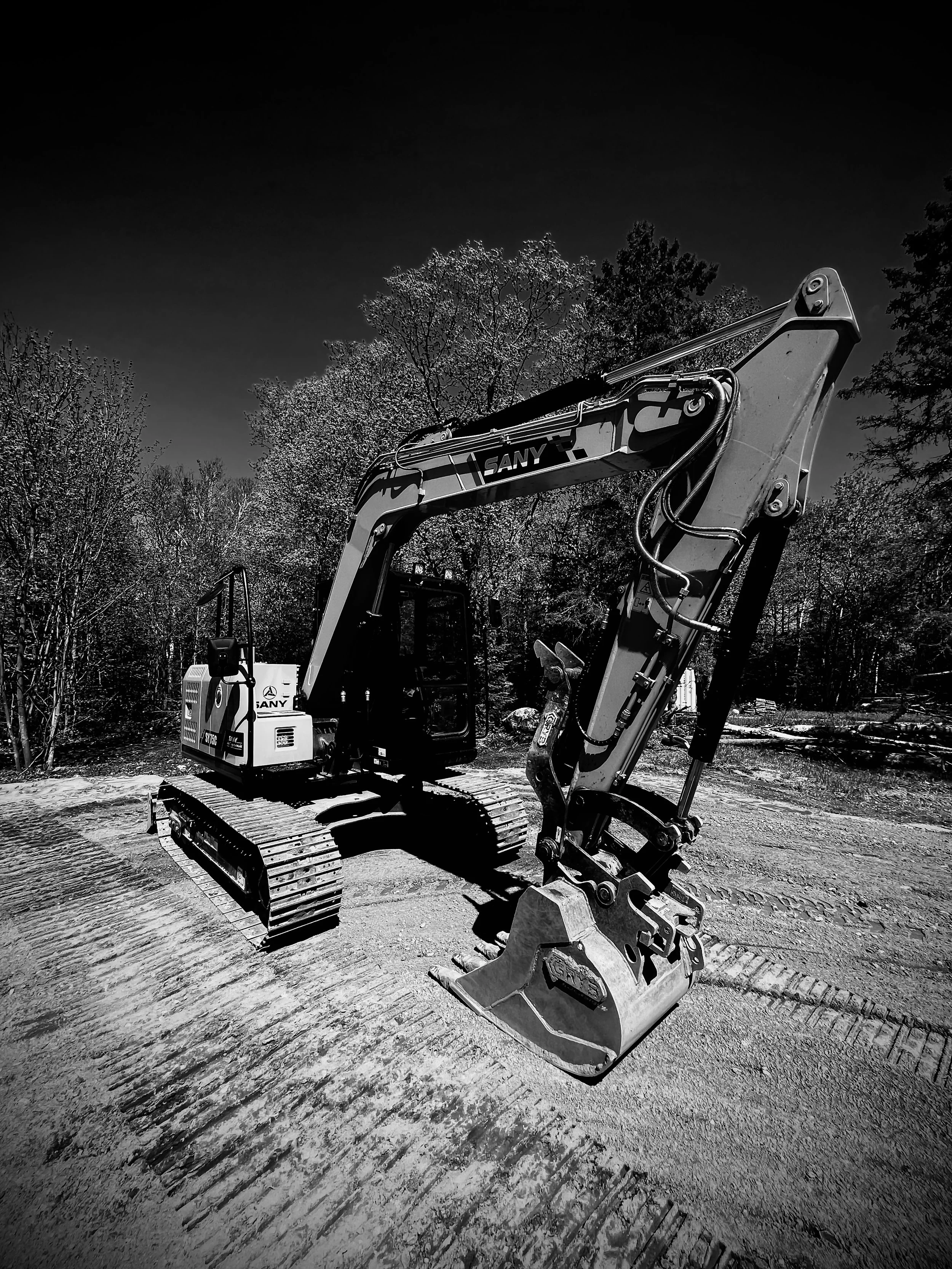 A black and white photo of a small SANY excavator on a construction site with trees in the background.