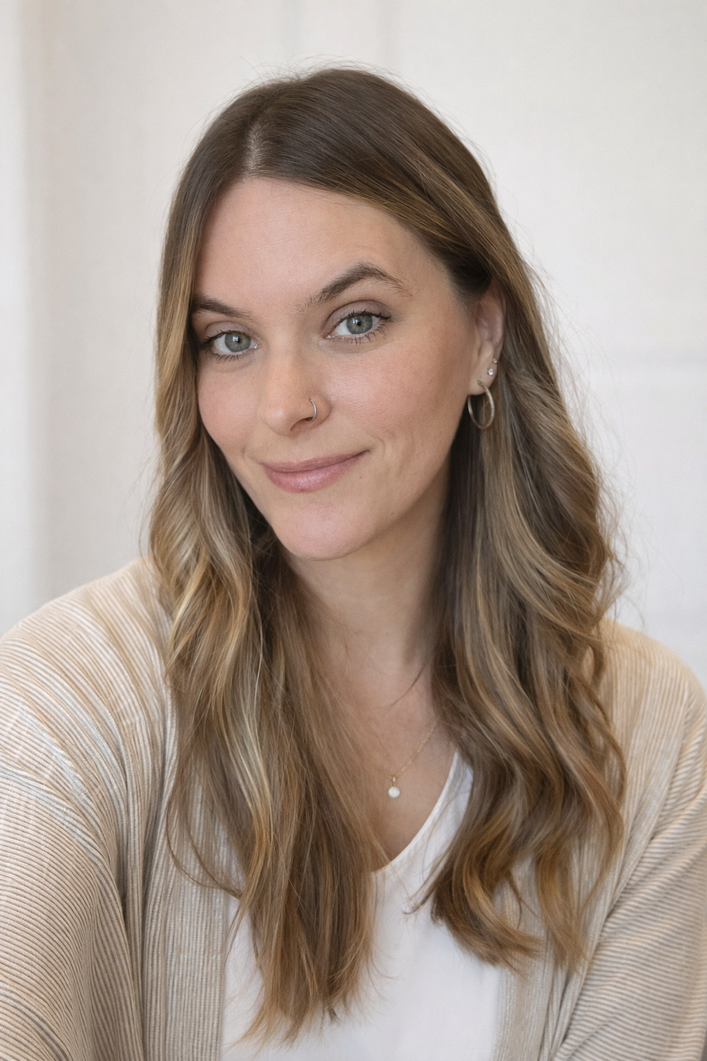 A woman with wavy, light brown hair and blue eyes, wearing a beige and white striped top, multiple earrings, and a small necklace, smiling softly.