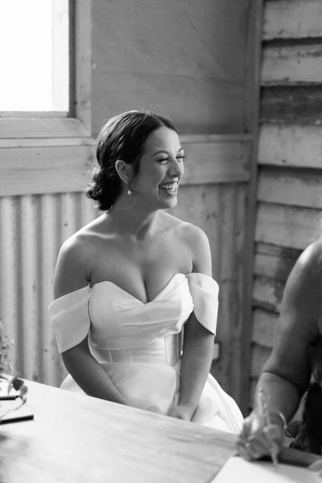 A woman in a white wedding dress with detachable sleeves, smiling and laughing, sitting at a table in a rustic wooden room.