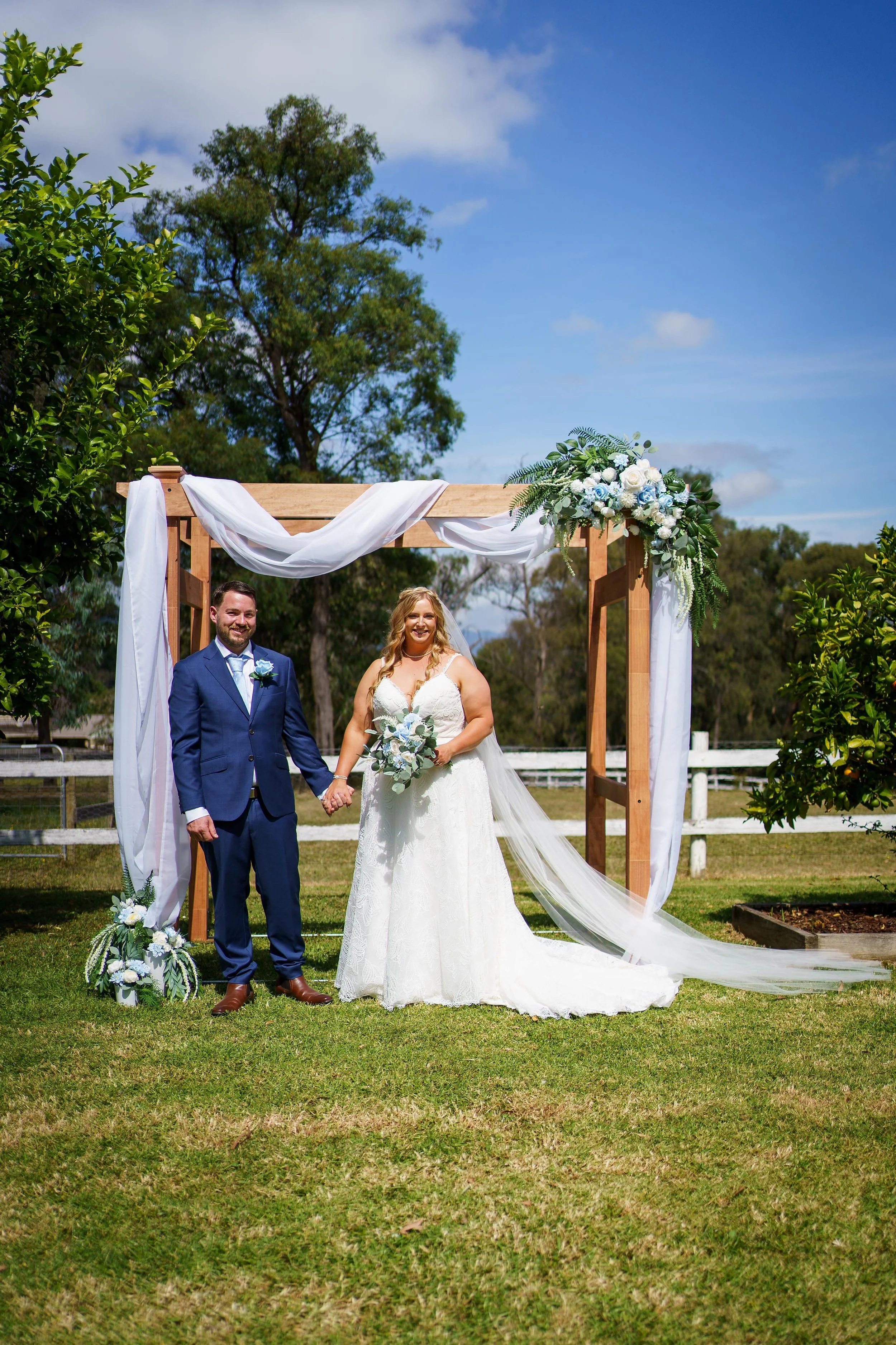 A bride and groom holding hands in front of a wooden wedding arch decorated with white fabric and flowers outdoors on a sunny day.