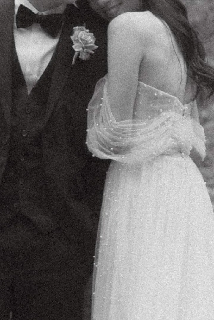 Black and white photo of a couple dressed in formal wedding attire, with the focus on the woman in a wedding dress and the man in a tuxedo with a bow tie and boutonniere.