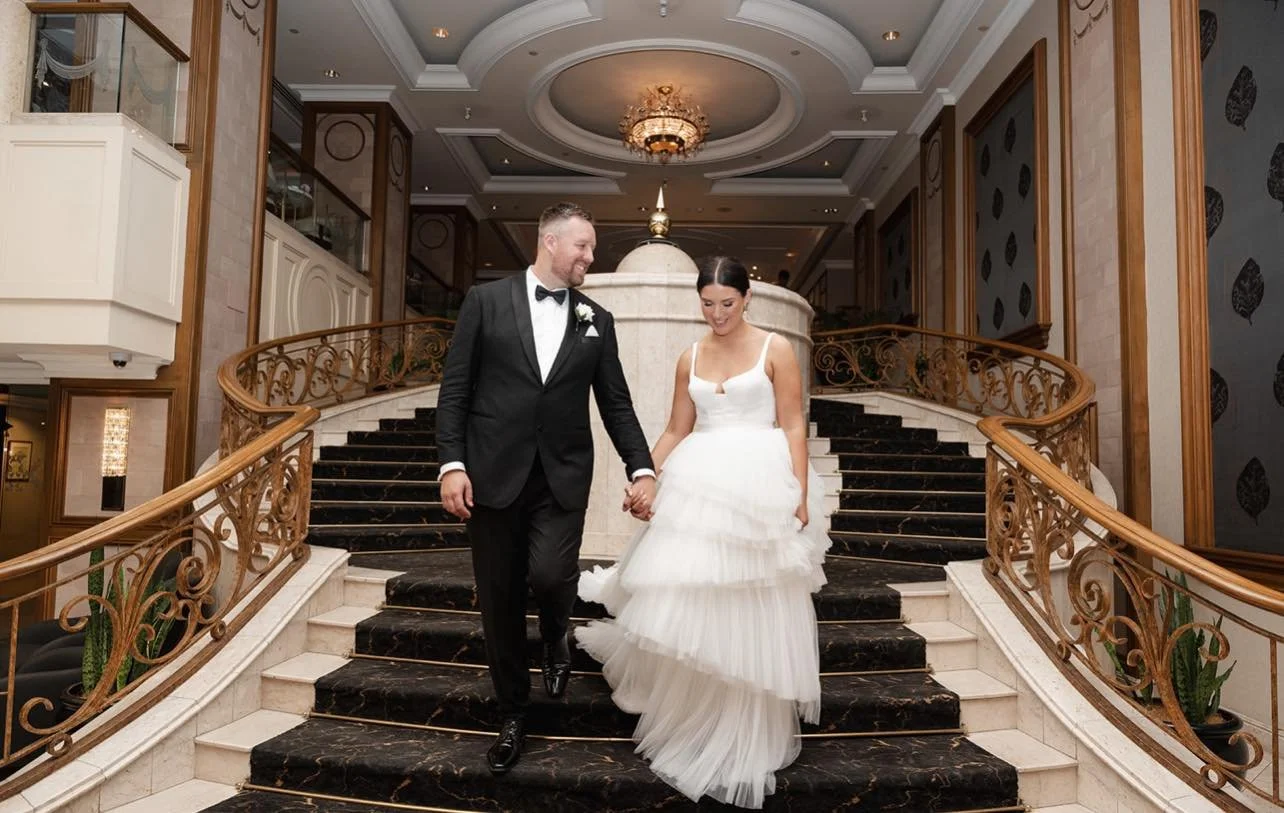 A bride and groom walk hand in hand down a grand staircase inside a decorated venue, with the bride wearing a white wedding gown and the groom in a black tuxedo.