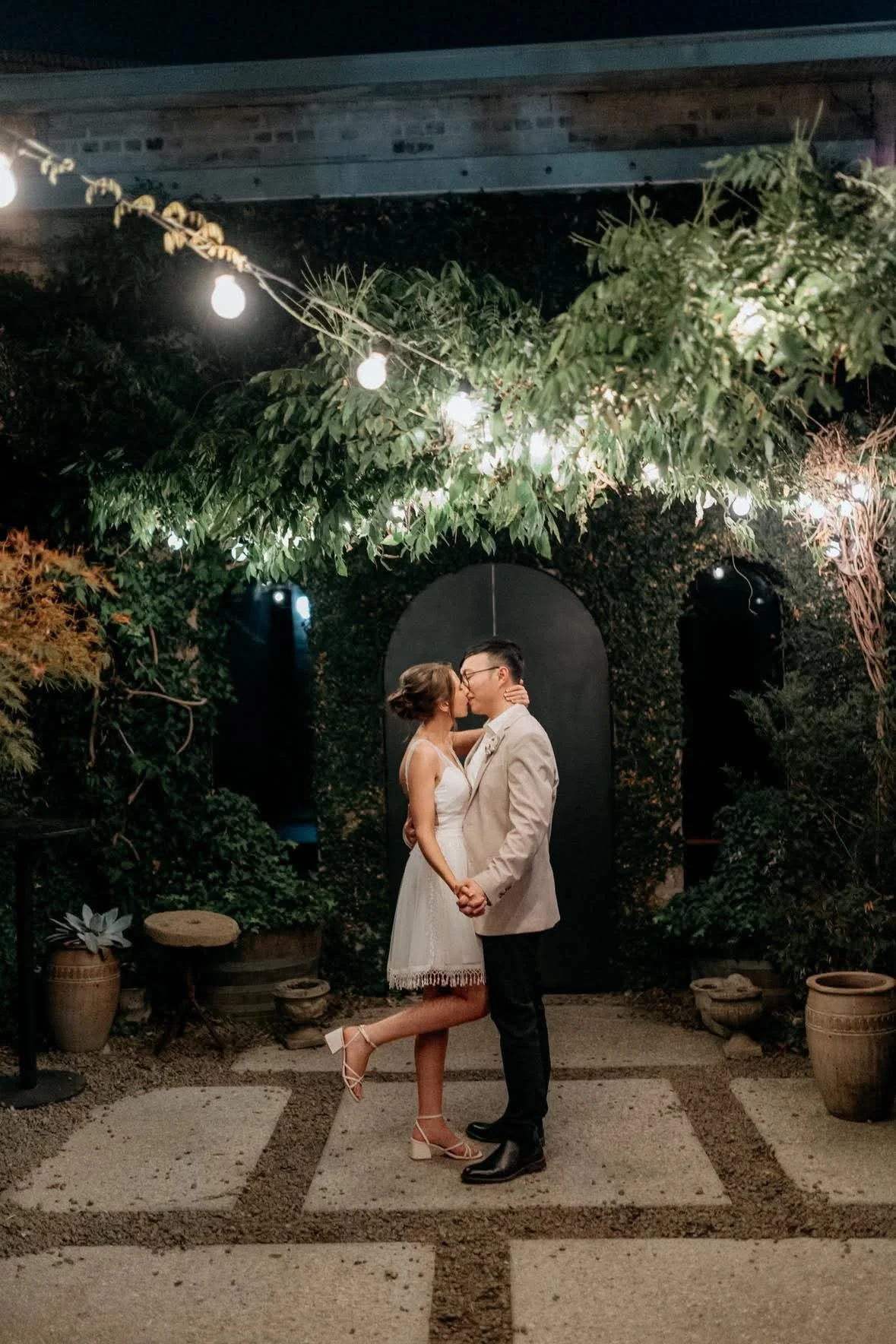 A newlywed couple sharing a kiss and holding hands in an outdoor garden at night, illuminated by hanging string lights and surrounded by lush greenery.