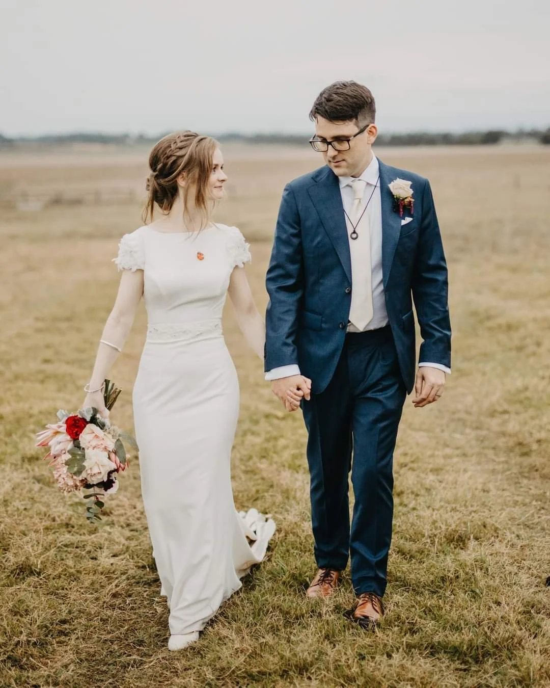 A bride and groom walking hand-in-hand on a grassy field, with the bride holding a bouquet of flowers and wearing a white wedding dress, and the groom in a blue suit with a boutonniere, during an outdoor wedding ceremony.