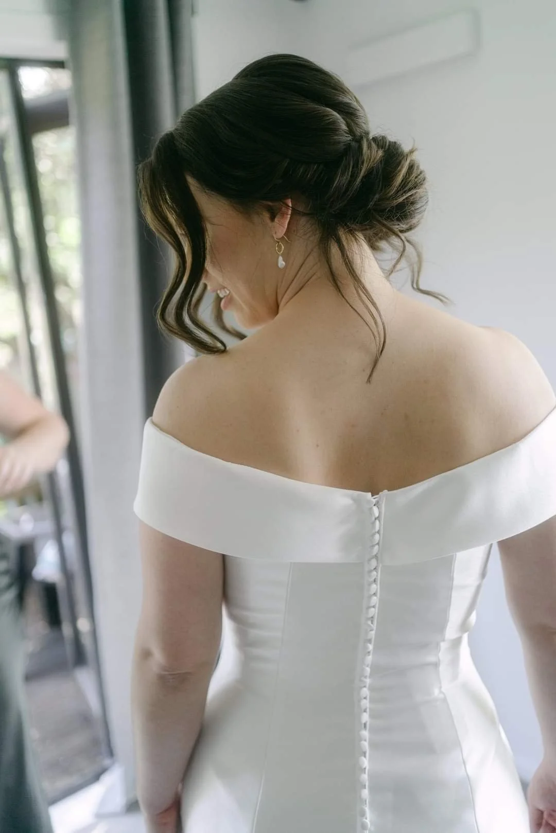 Rear view of a woman in a white off-shoulder dress, with dark wavy hair styled in an updo.