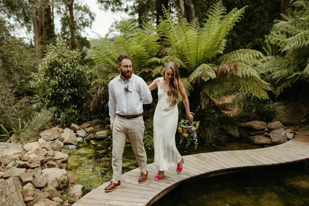 A couple walking on a curved wooden footbridge over a pond in a lush garden with tall ferns and greenery.