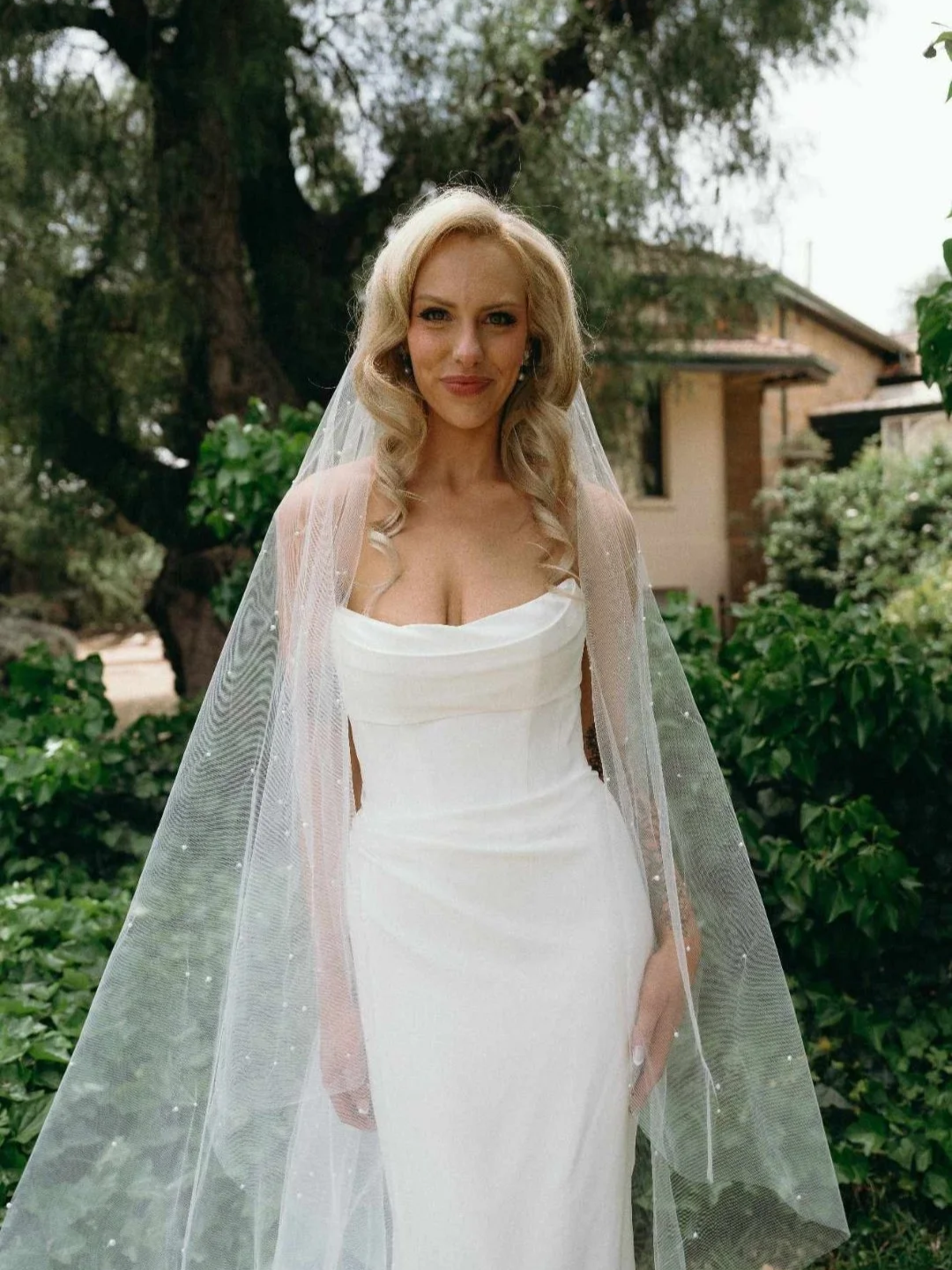 A woman in a white wedding dress with a bespoke sheer veil standing outdoors among green foliage, with trees and a house visible in the background.