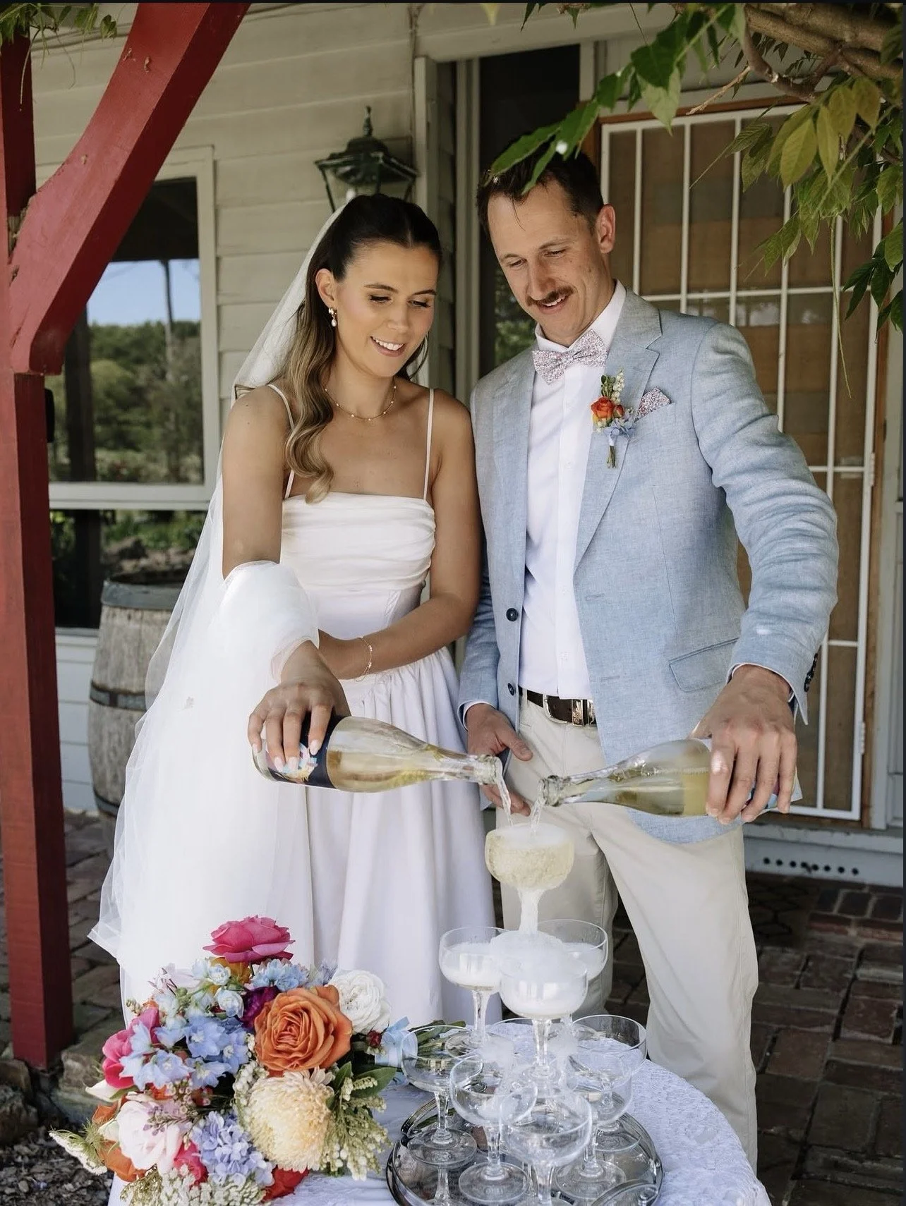 A bride and groom pour champagne into glasses on a table decorated with flowers.