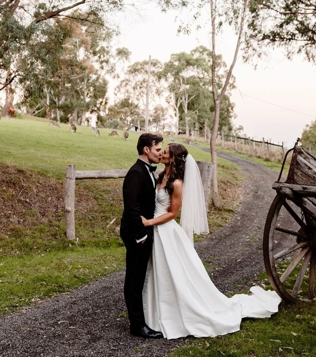 A bride and groom kissing outdoors on a dirt path, with a green hillside, trees, and a wagon wheel to the right in the background.