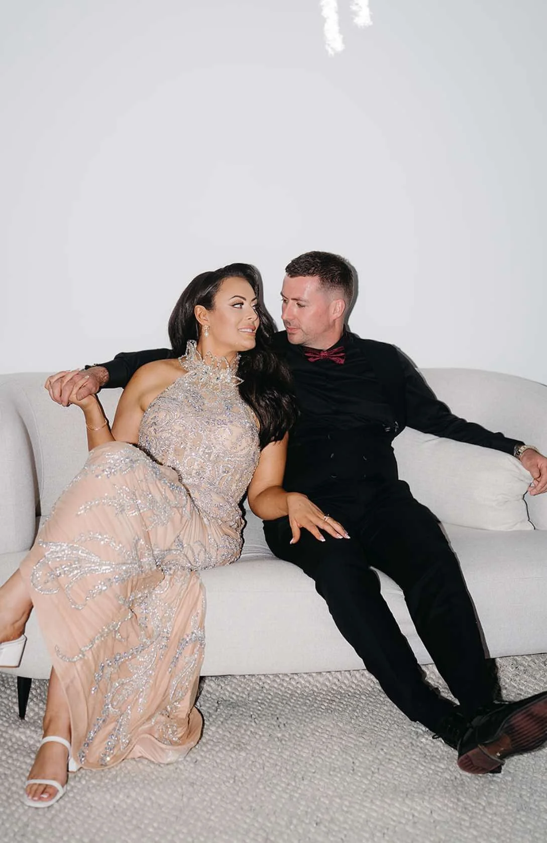 A couple sitting on a white couch, dressed in a beaded albina dyla dress, gazing at each other lovingly.