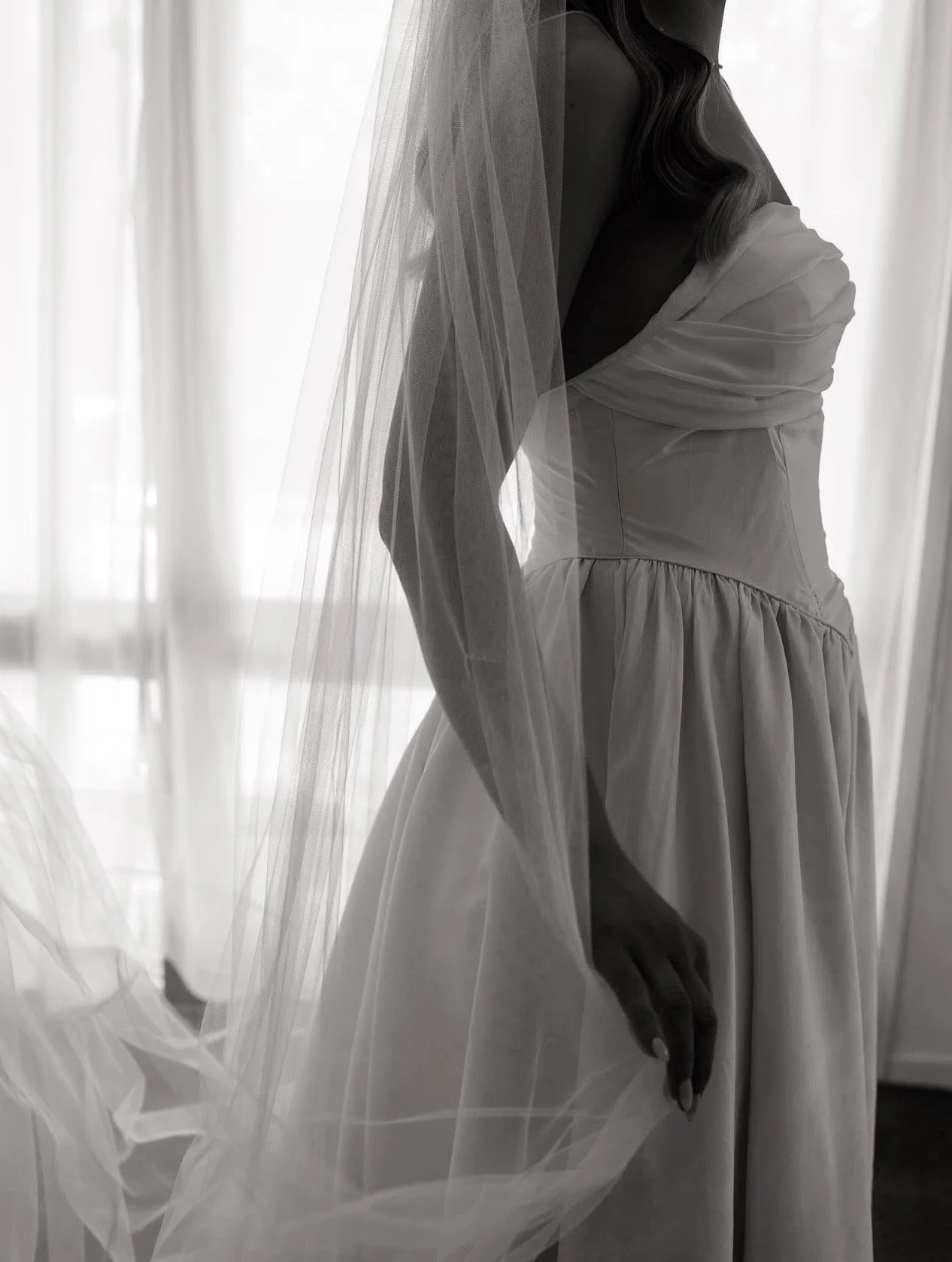A woman in her mums wedding dress with a custom made veil, standing near a window with sheer curtains, in black and white.