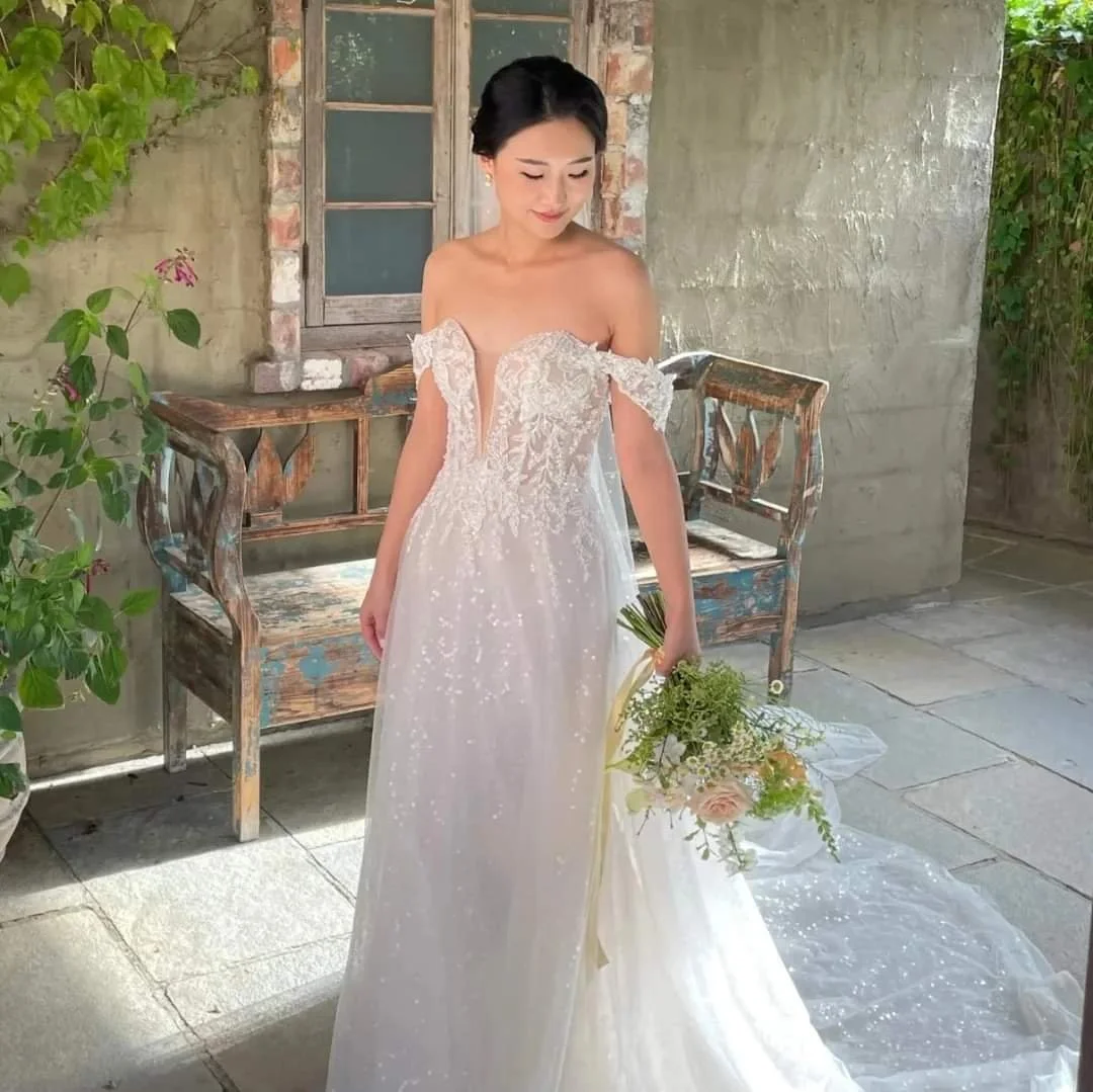 A woman in a white wedding dress holding a bouquet of flowers, standing outdoors on a tiled patio next to a weathered wooden bench and a plant, in front of a brick and concrete wall.