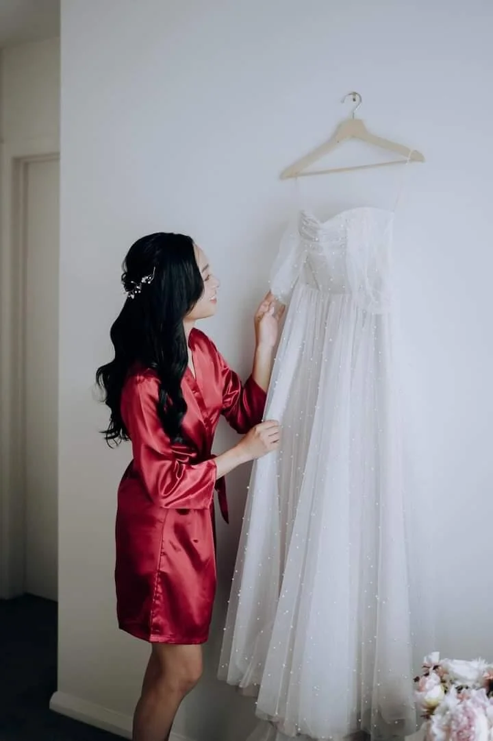 A woman with black hair in a red satin robe looking at a white wedding dress hanging on a wall hook.
