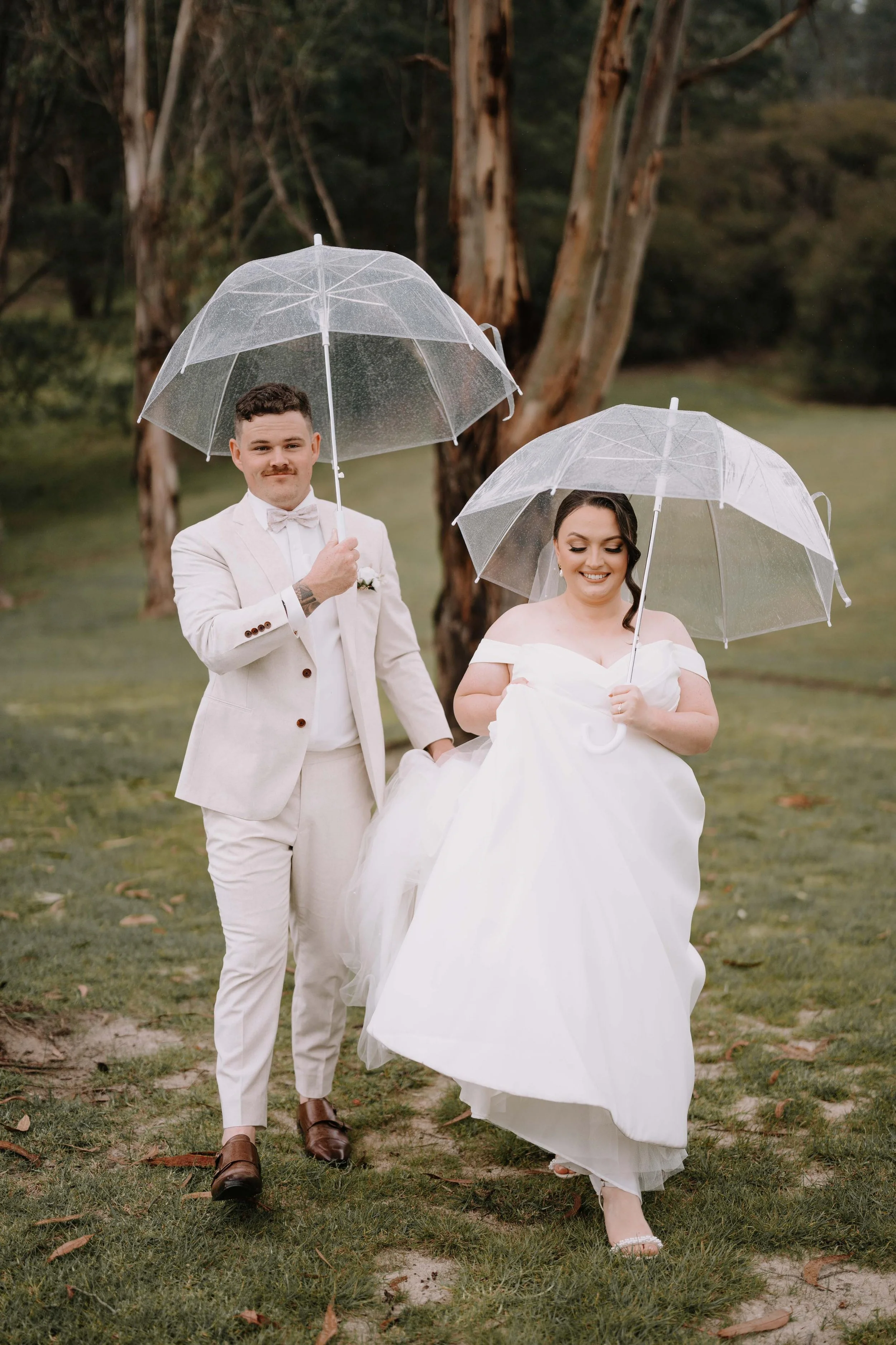 A bride and groom in wedding attire holding umbrellas outdoors on a rainy day.