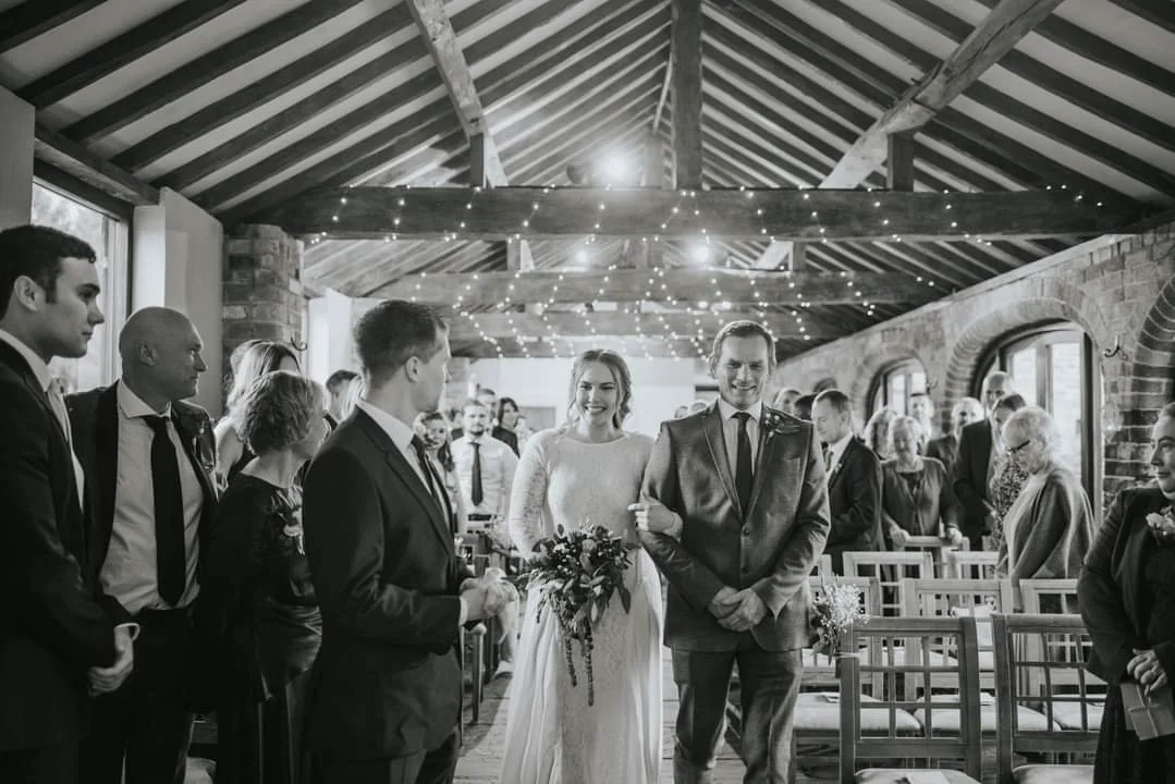 A wedding ceremony with a bride and groom walking down the aisle, surrounded by guests in an indoor venue with string lights and brick walls.