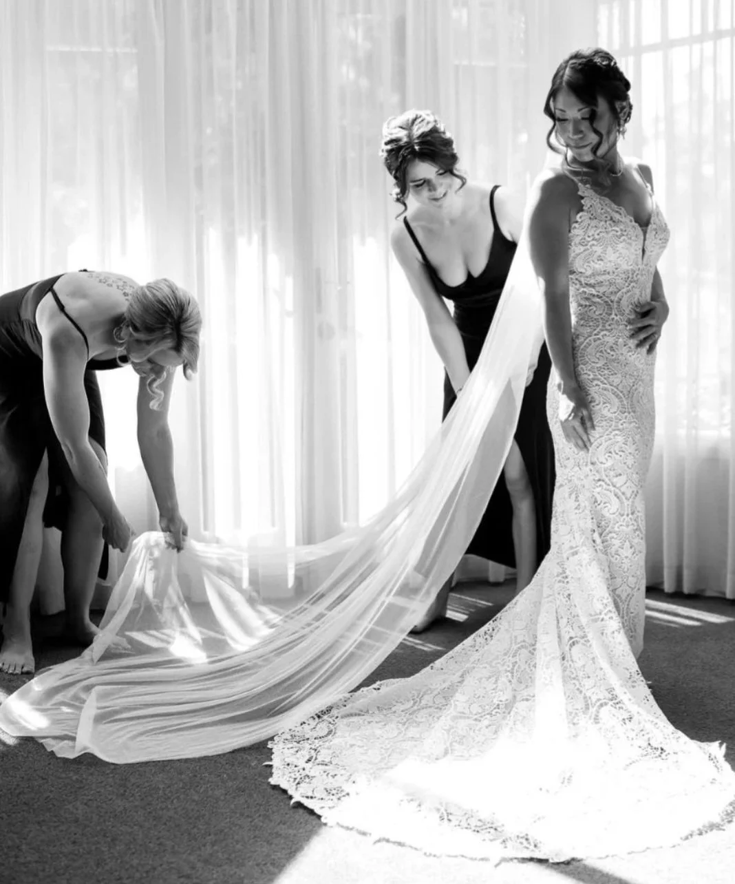 Four women, one in a wedding dress and three in dark dresses, are adjusting the bride's long lace train in a room with sheer curtains, sunlight streaming in.