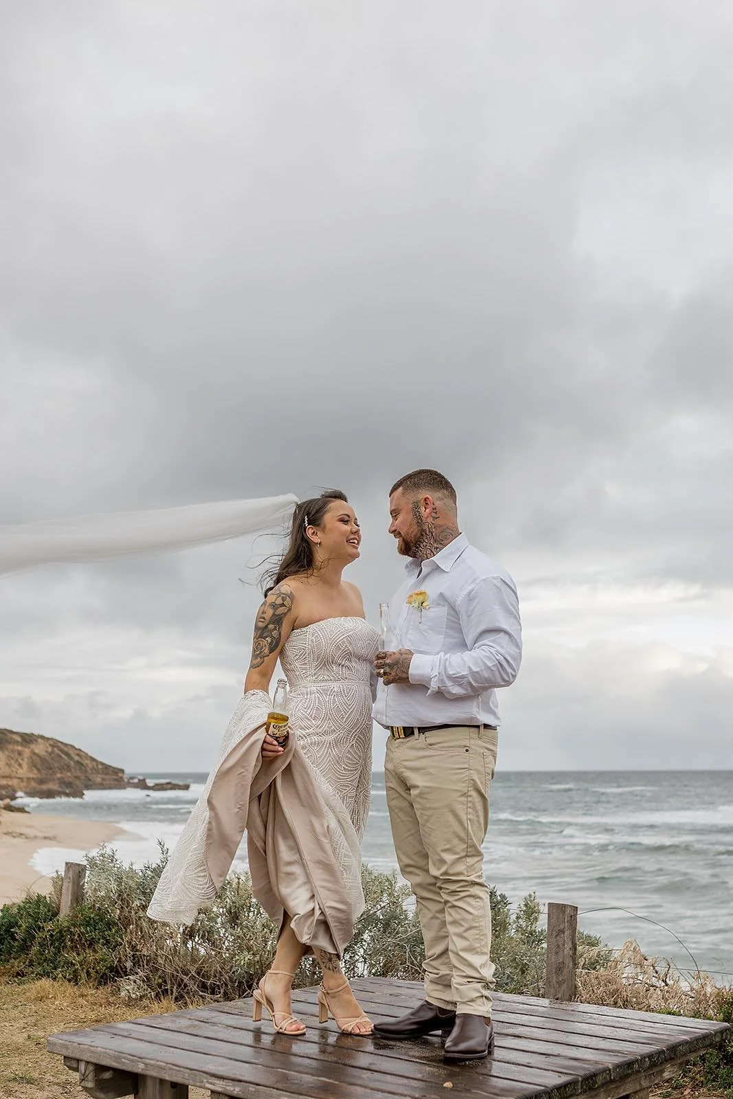 A bride and groom stand on a wooden platform by the beach, smiling and holding drinks, with overcast skies and ocean waves in the background.