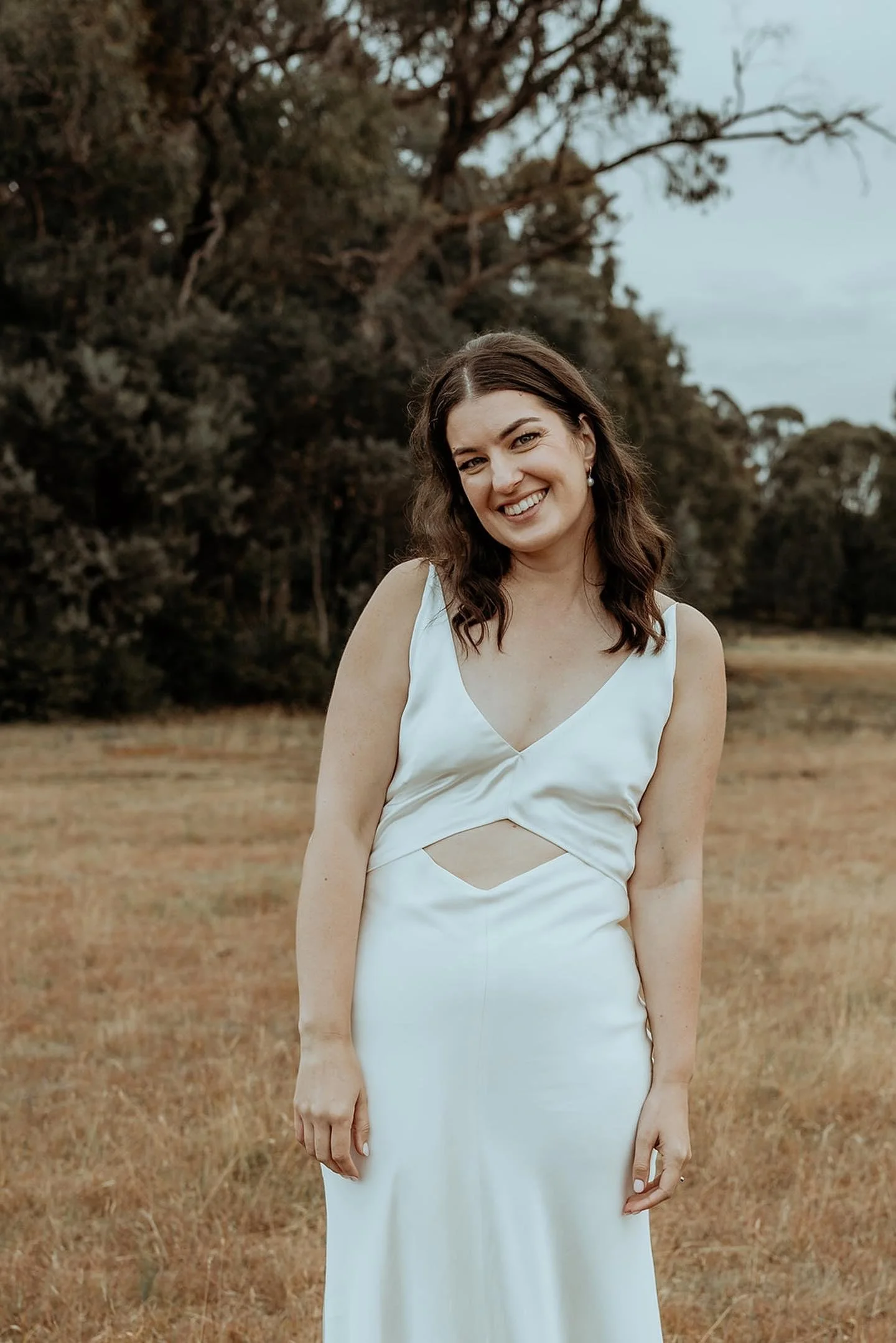 A woman in a white sleeveless dress standing outdoors in a grassy field with trees in the background, smiling at the camera.