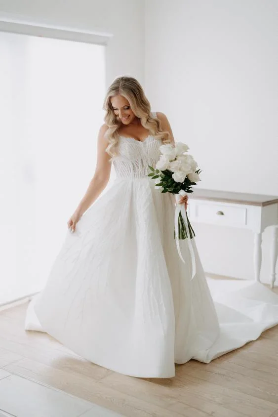 Bride in a beaded white wedding gown holding a bouquet of white roses, standing in a bright room after expert bridal alterations.