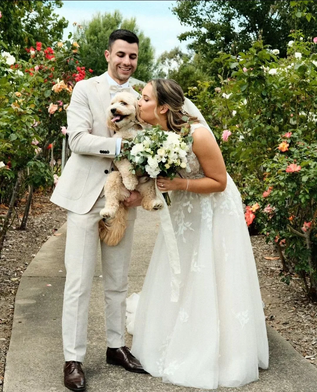 Bride and groom in wedding attire sharing a tender moment with their golden retriever dog, outdoors in a garden with blooming flowers, the bride holding a bouquet of white flowers.