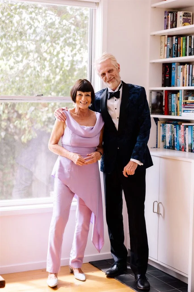 An elderly woman and man in formal attire standing beside each other, smiling, in a brightly lit room with a window and bookshelf.