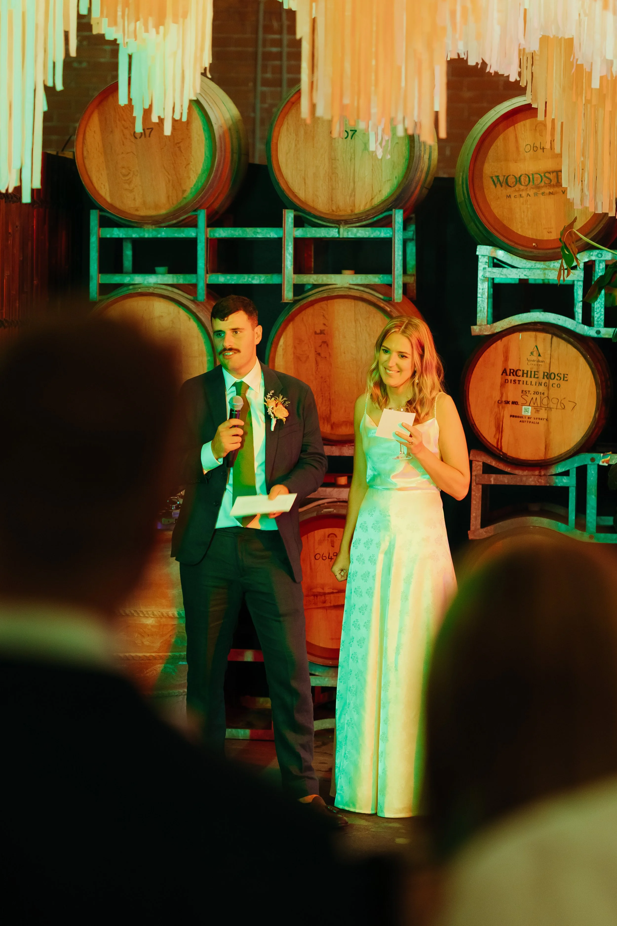 A man and a woman giving a speech at a wedding reception, standing in front of large wine barrels in a rustic setting.