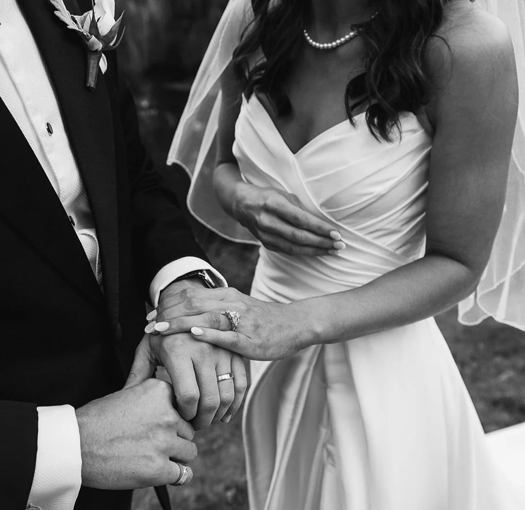 A black and white photo of a bride and groom holding hands. The bride is wearing a wedding dress and pearl necklace, and the groom is in a tuxedo with a boutonniere. Their wedding rings are visible.